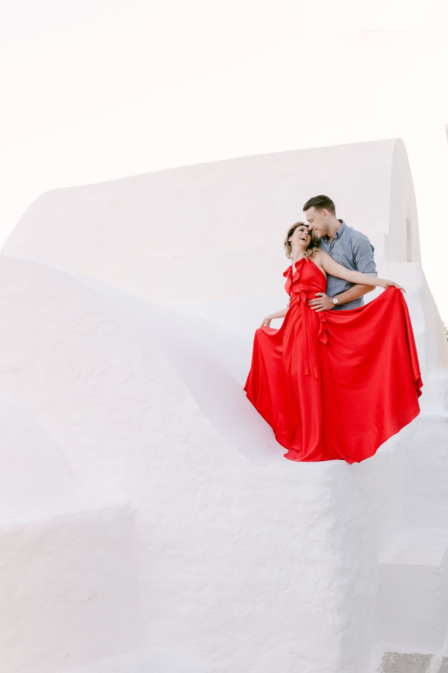 Elegant couple embracing on white wall with flowing red dress.