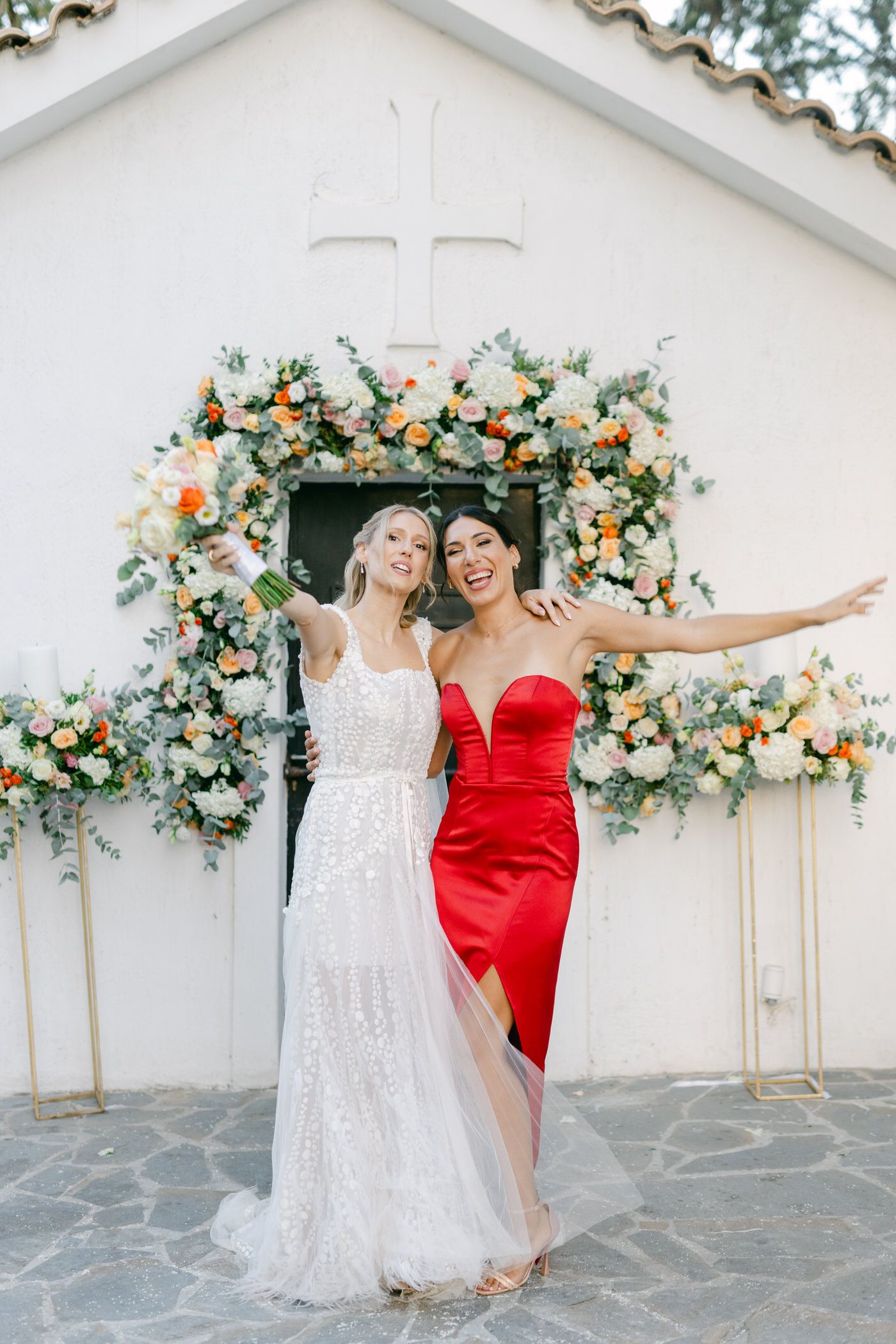Two women in formal dresses pose together under a floral arch by a white church entrance with a cross above.