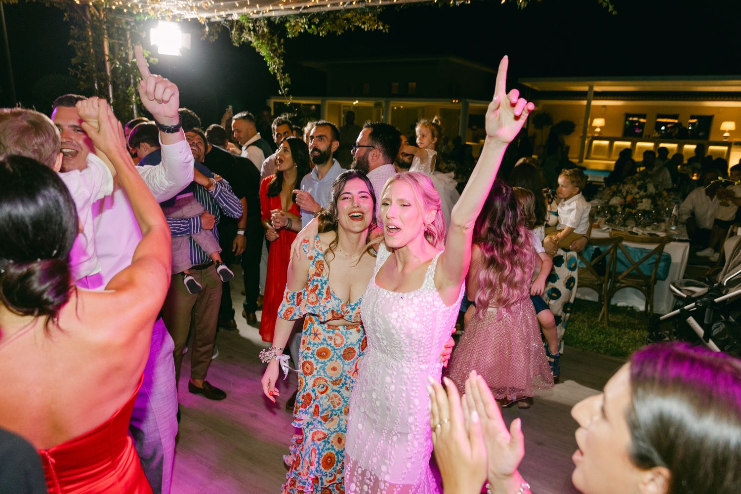Wedding guests dance and celebrate on a lively dance floor illuminated by pink and purple lighting at an evening reception.