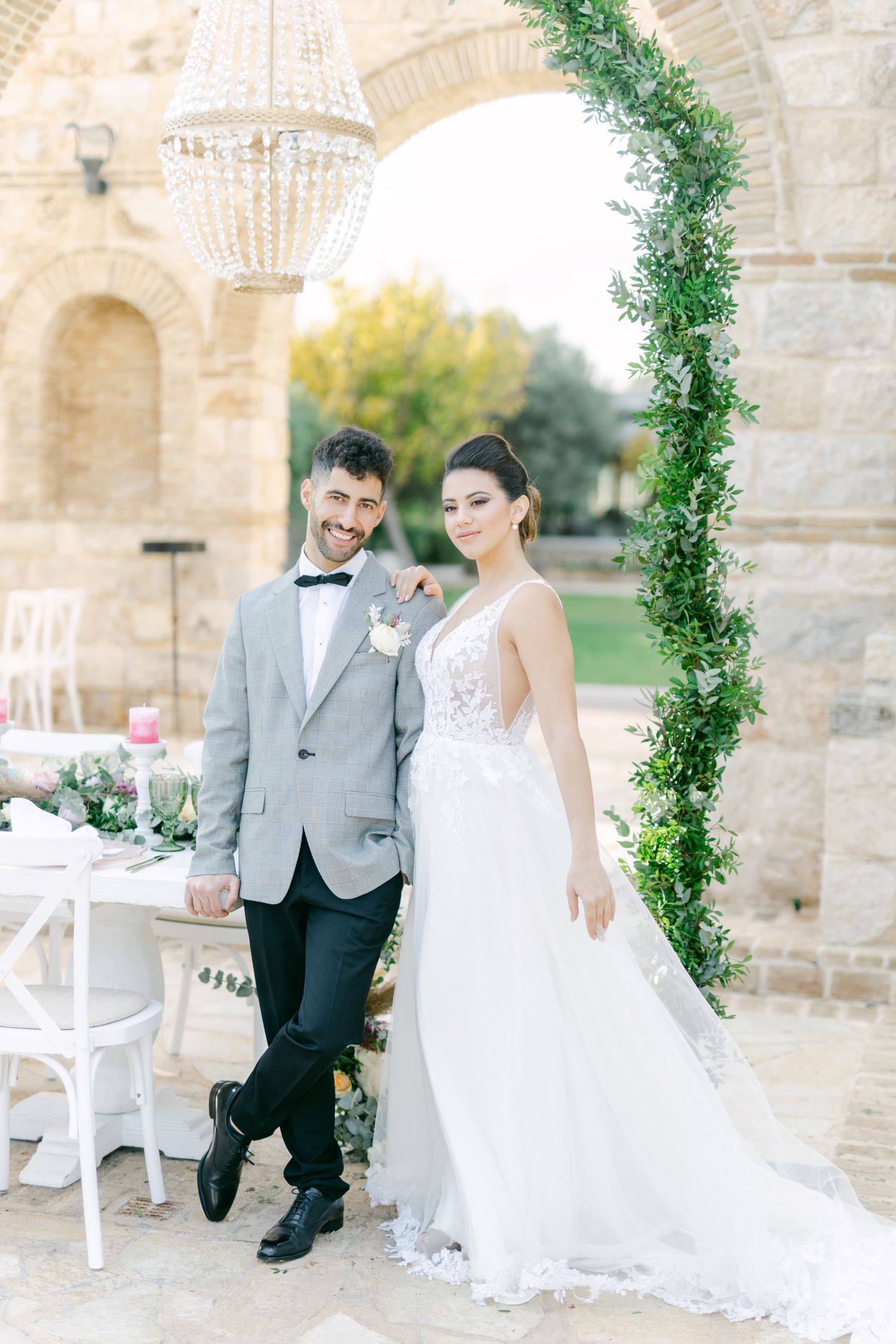 Wedding couple poses under stone archway decorated with greenery and crystal chandelier.