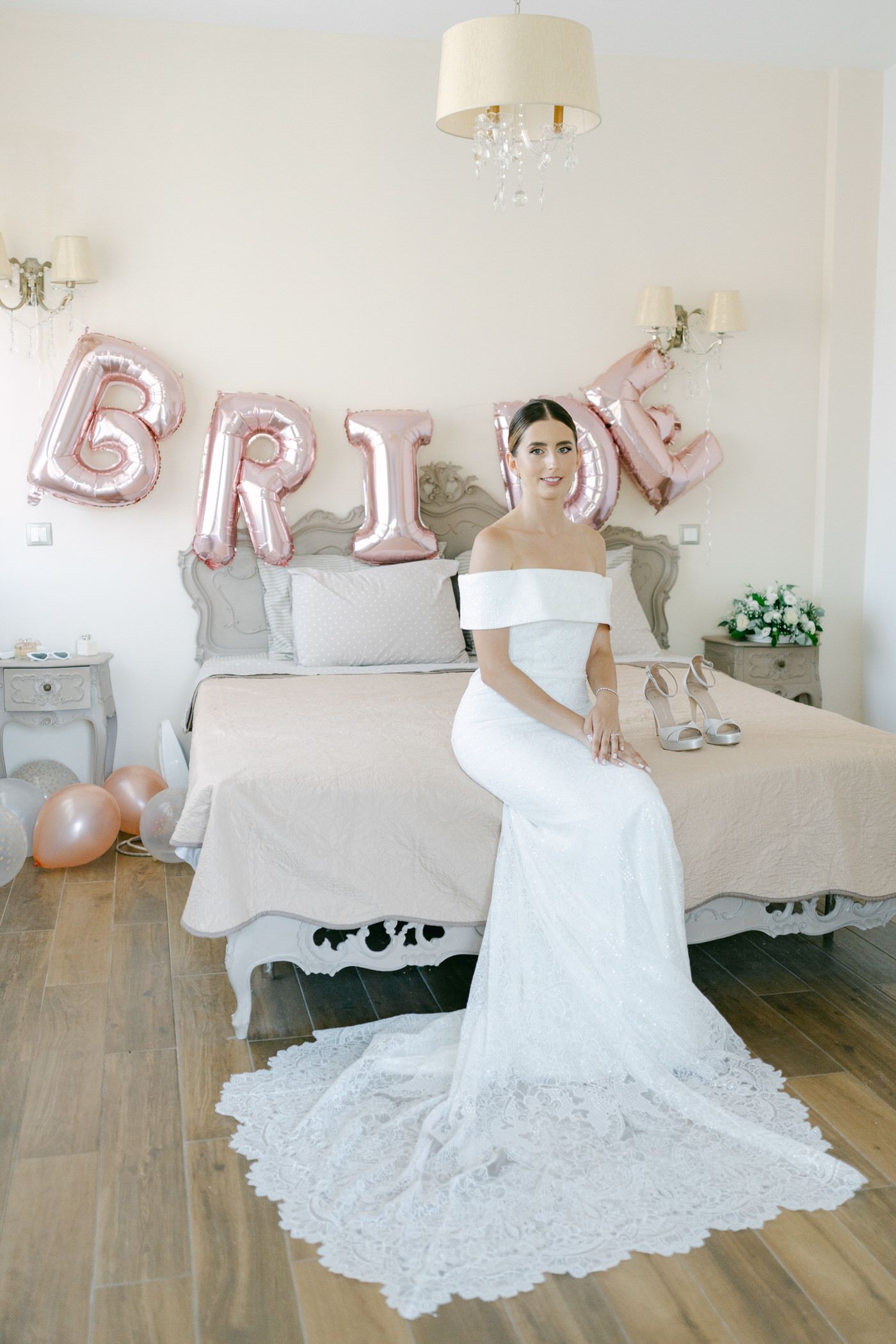 A white lace wedding dress with off-shoulder design displayed in a bedroom decorated with rose gold 'BRIDE' balloons.