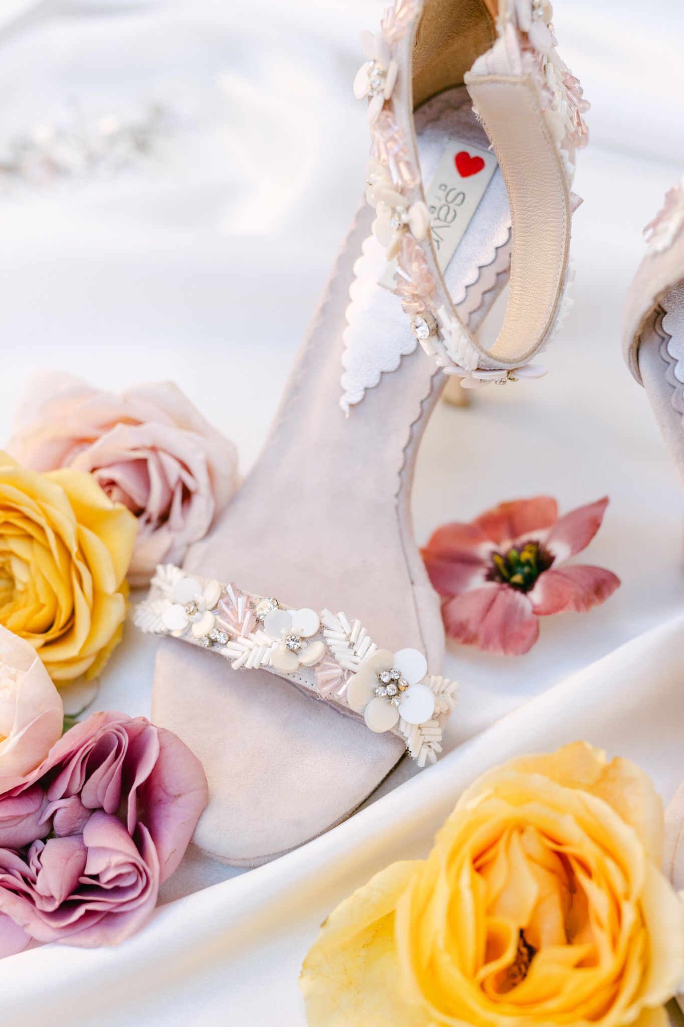 Elegant white bridal shoes with lace detail surrounded by yellow and pink roses on white fabric background.
