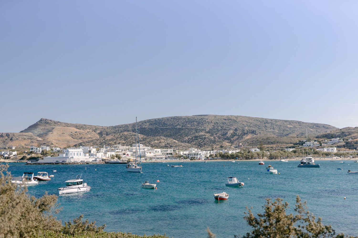Boats anchored in turquoise Mediterranean waters near coastal Greek town with hillside landscape in background.