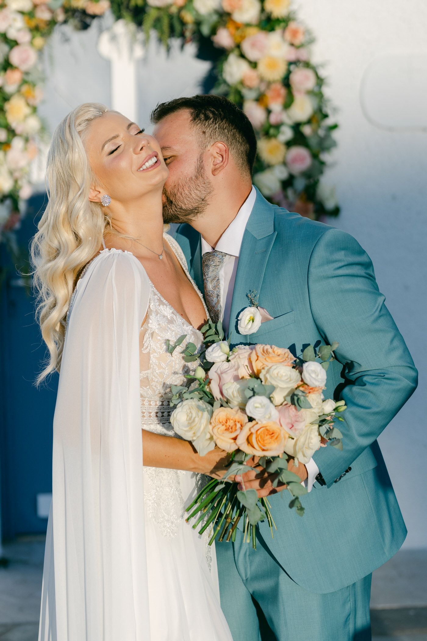 A romantic wedding moment under a floral arch with peach and white roses, the couple holding a beautiful bouquet.