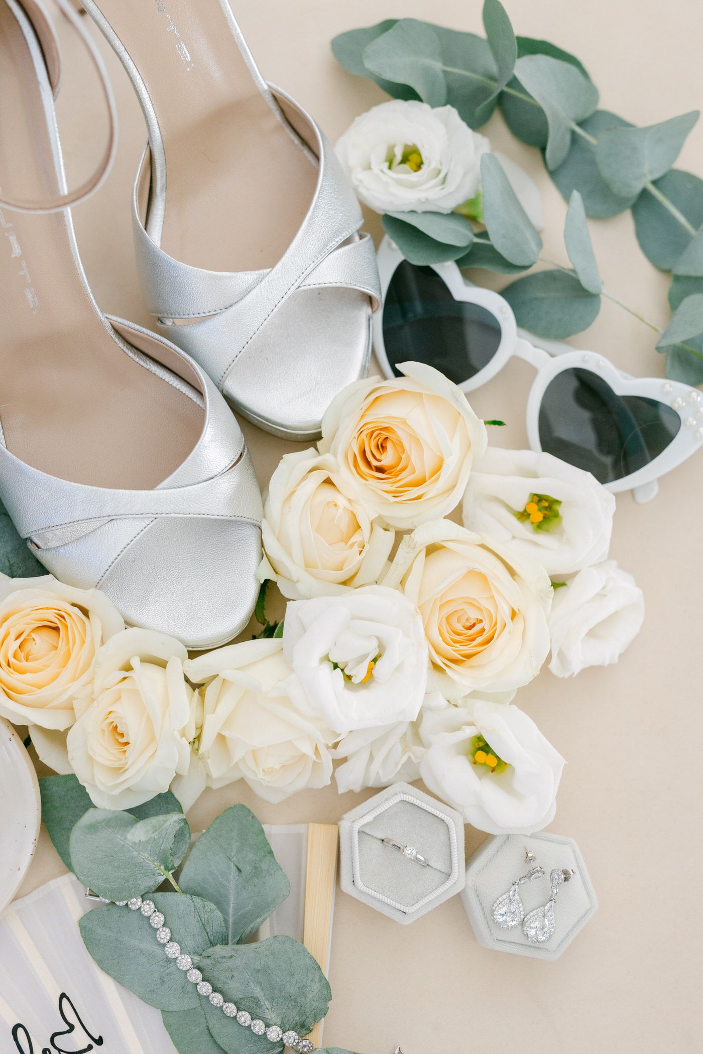 White bridal shoes with elegant roses, eucalyptus leaves and ring boxes arranged in a romantic wedding flatlay.