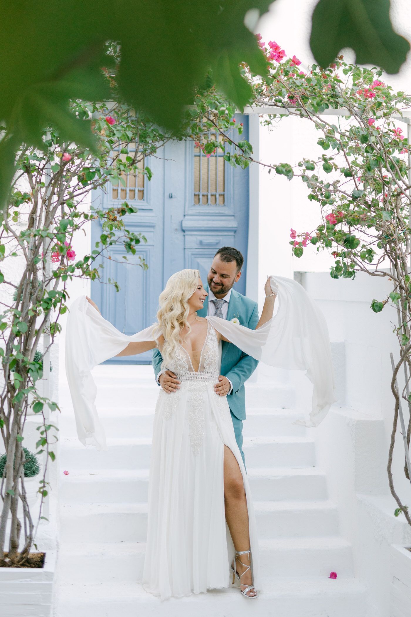 A romantic wedding couple posing on white stairs with pink flowers and a blue door in Mediterranean style setting.
