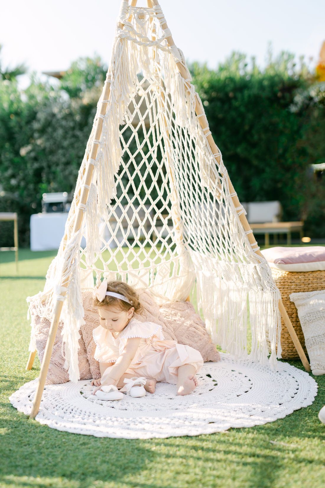 A decorative macrame teepee tent with a white crochet rug sits on a lawn in a garden setting.