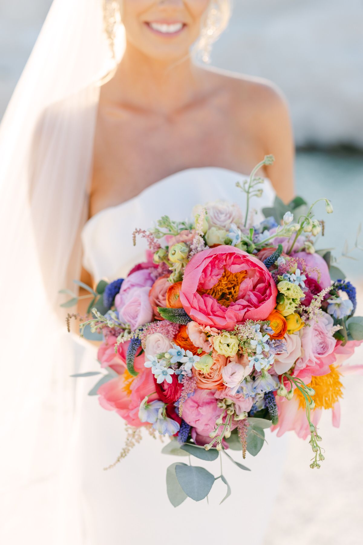 Vibrant wedding bouquet with pink peonies, roses and mixed flowers in coral and purple tones against white dress.
