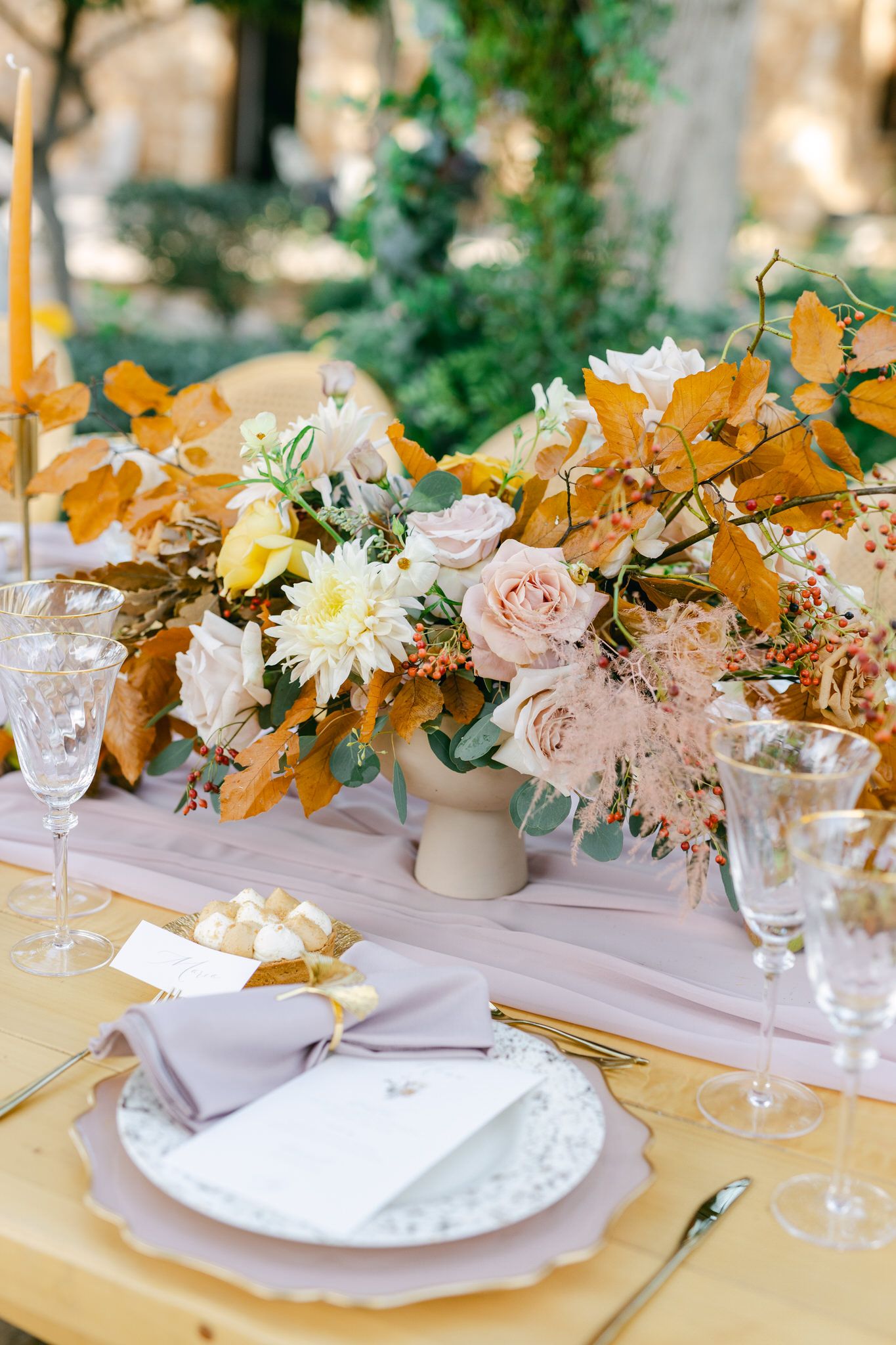 Elegant table setting with blush roses and golden fall foliage centerpiece on white linens and yellow tablecloth.