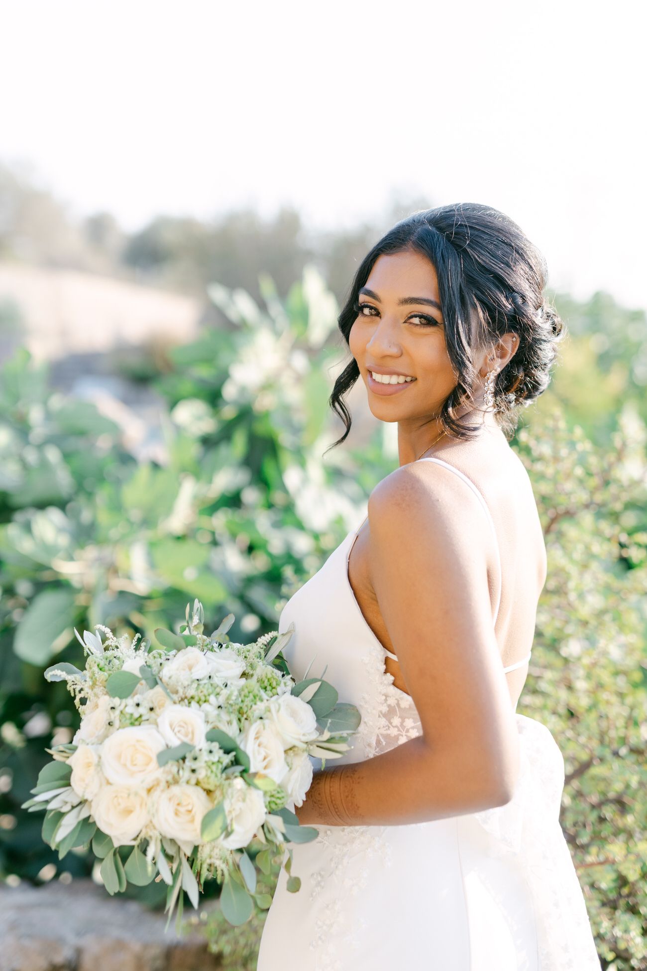 Bride in white dress holding bouquet of white roses and greenery against sunlit garden background.