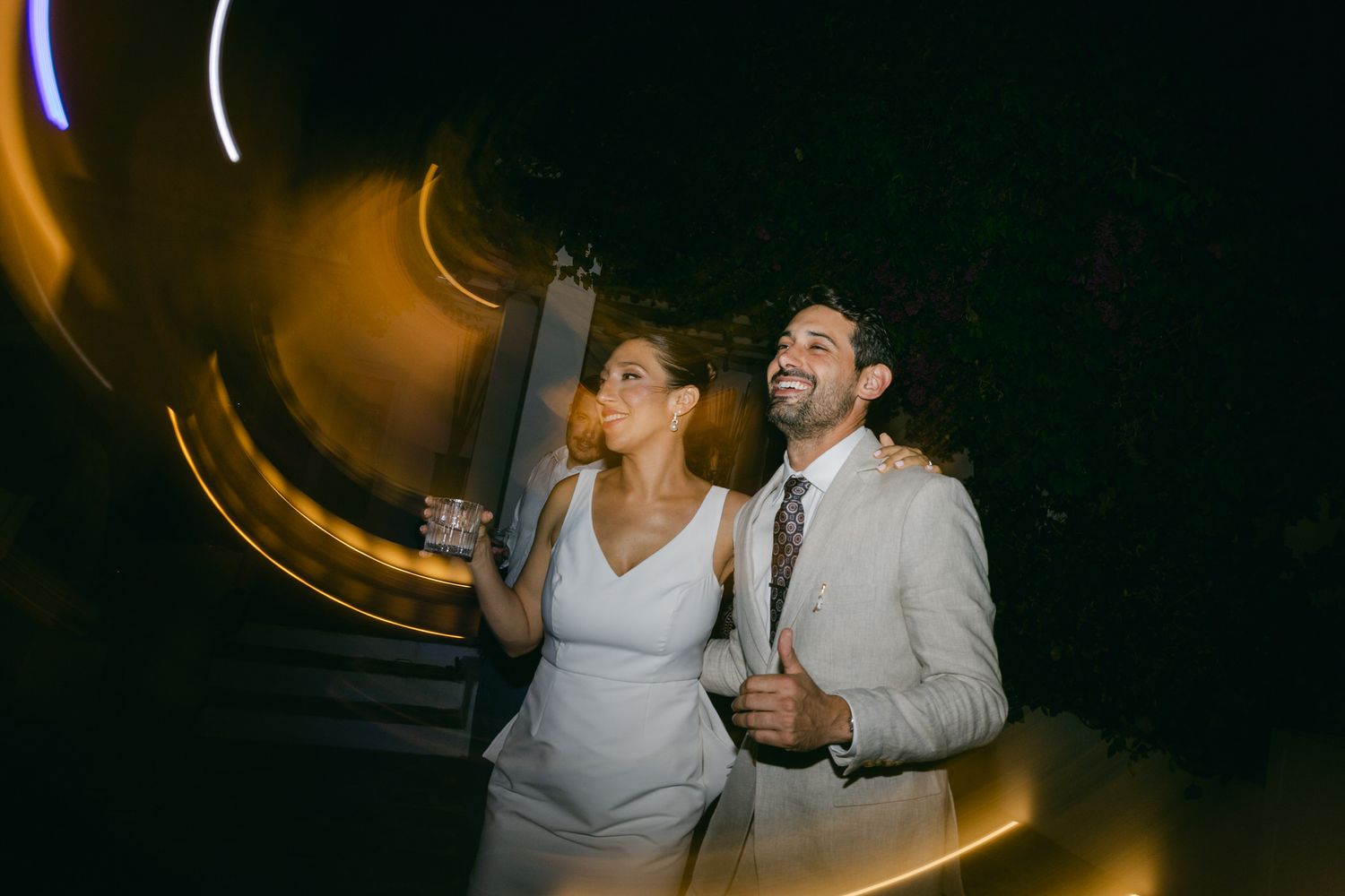 A couple in wedding attire dance and laugh together at a darkly lit reception with glowing light trails in the background.