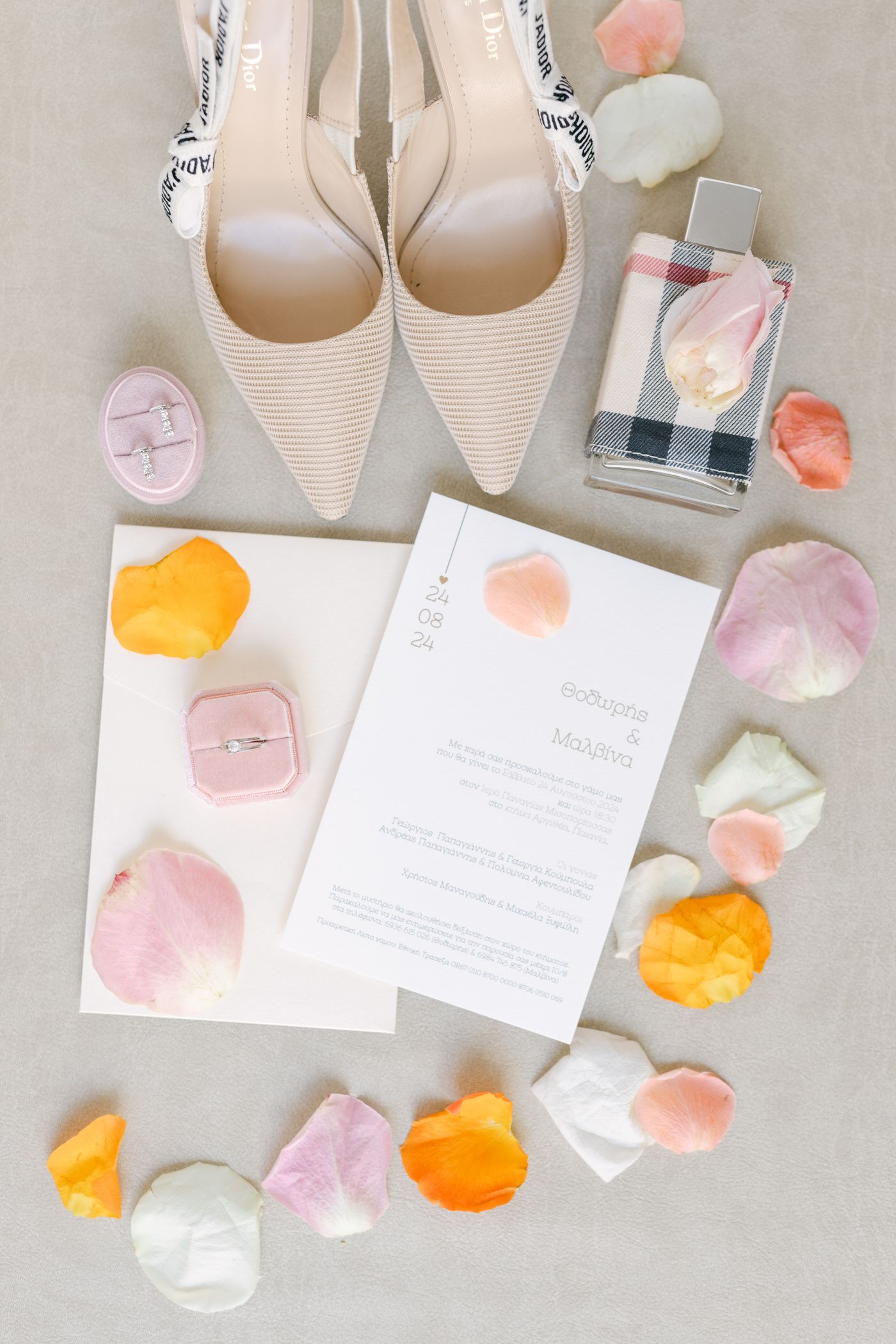 A wedding invitation flatlay with elegant beige heels, rose petals in pink and orange, and a minimalist white invitation card.