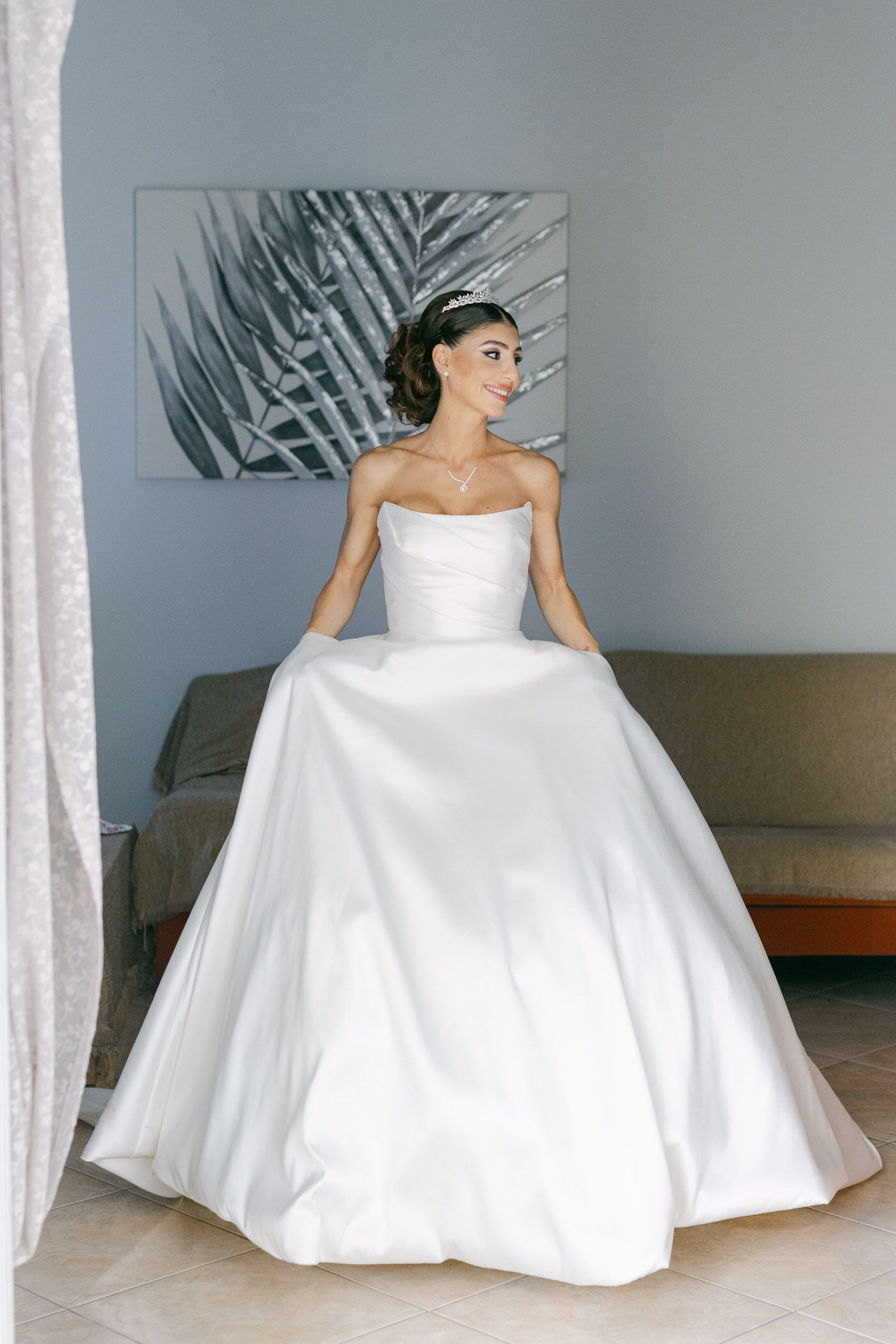 A person in a stunning white strapless ballgown wedding dress poses in front of a palm leaf artwork on a gray wall.