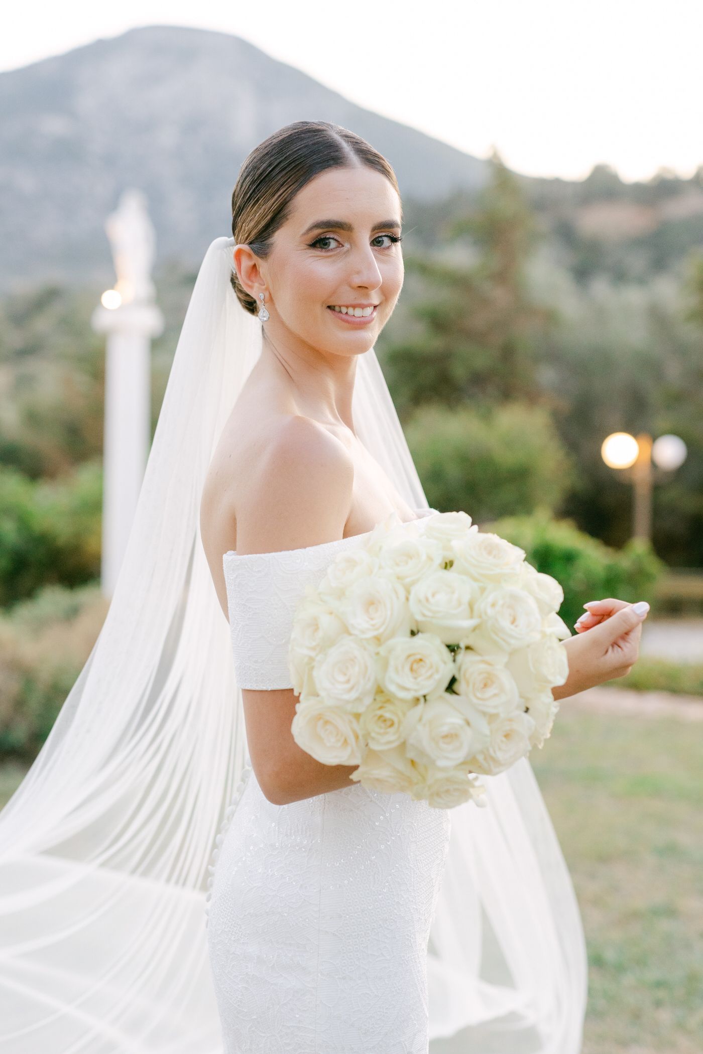 A bride in an elegant off-shoulder white wedding dress holds a luxurious bouquet of white roses with a flowing veil.