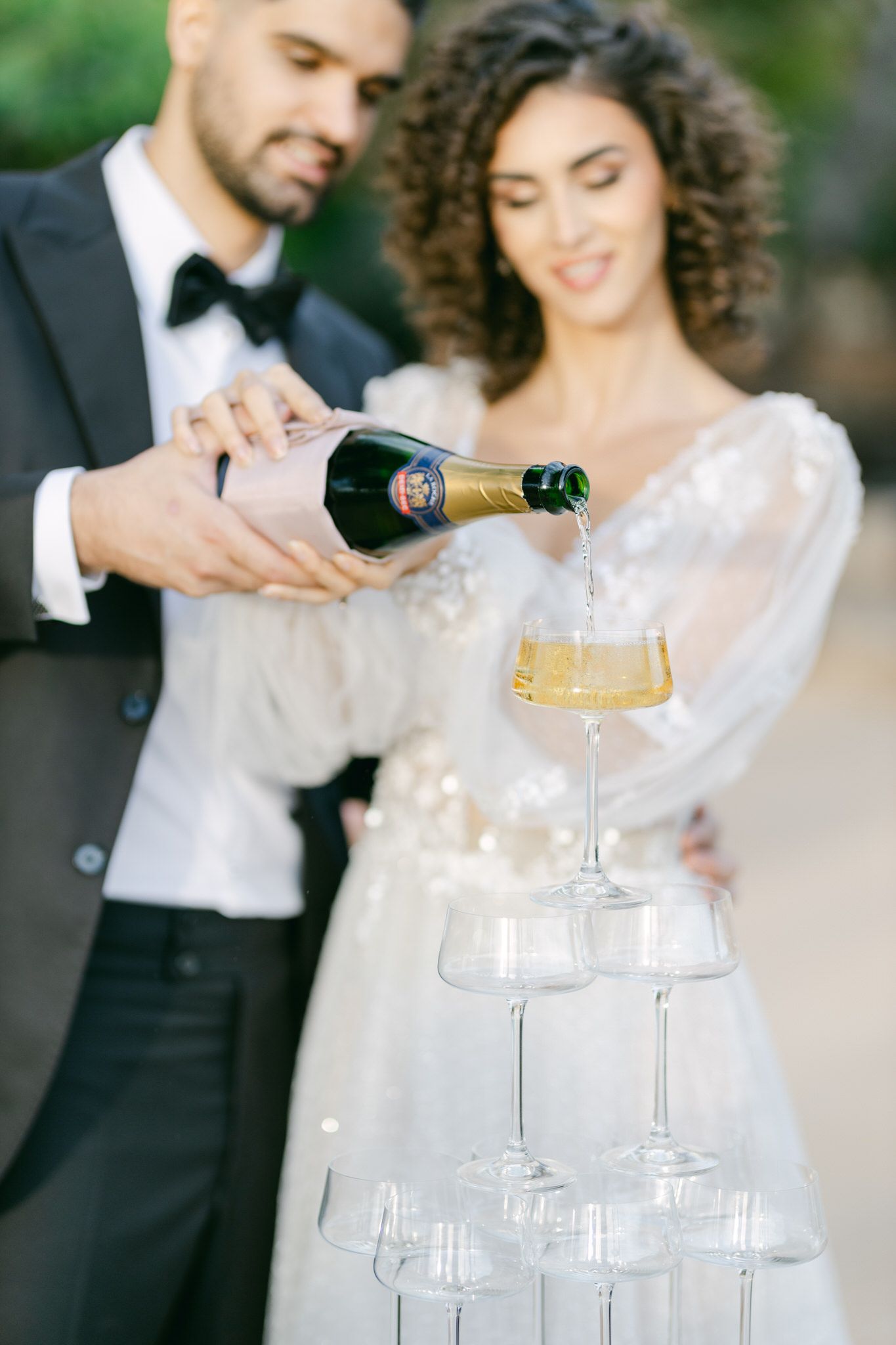 A couple pours champagne into a tower of stacked crystal glasses at an elegant wedding celebration.