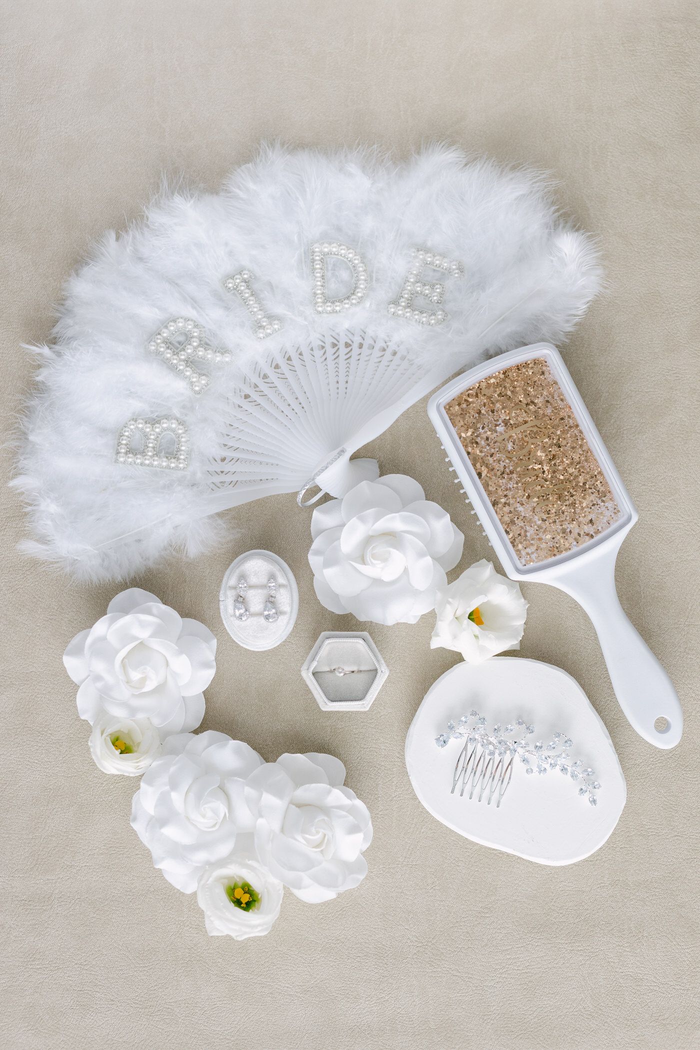 White decorative bridal accessories including feather fan, floral accents, jewelry, and glitter details on beige background.