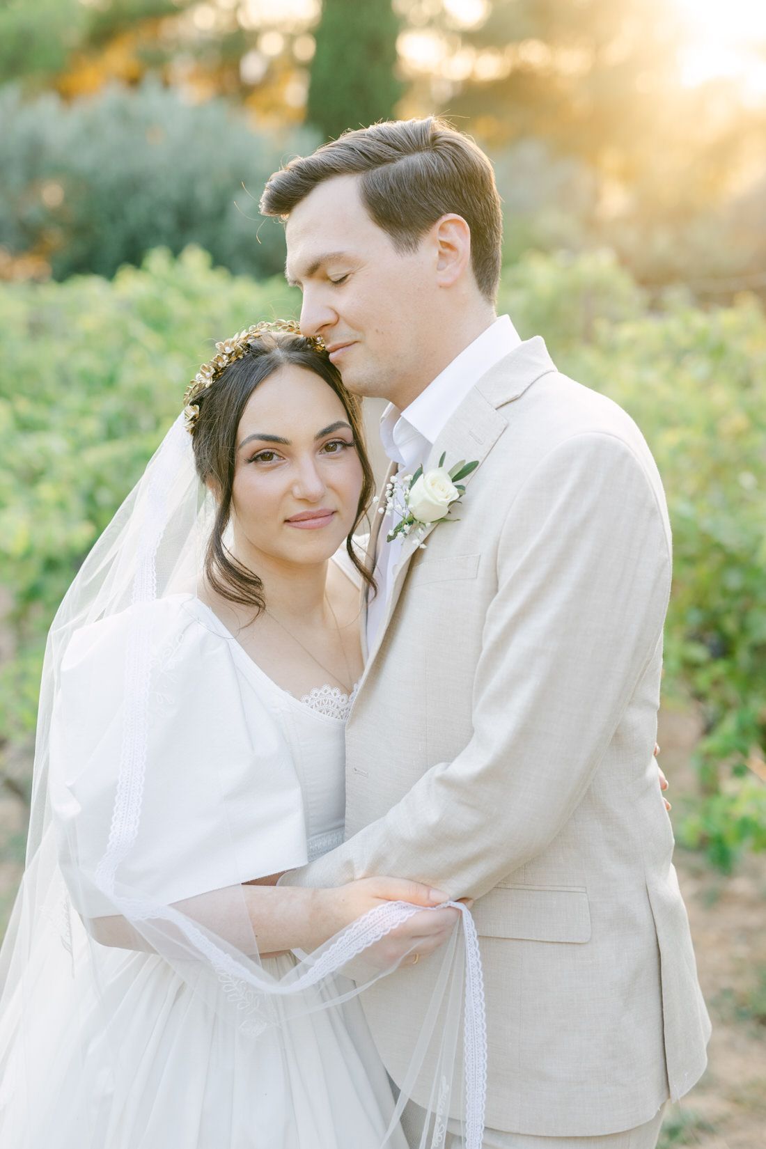 A romantic wedding couple embraces in a vineyard during golden hour, dressed in white and beige attire with delicate floral details.