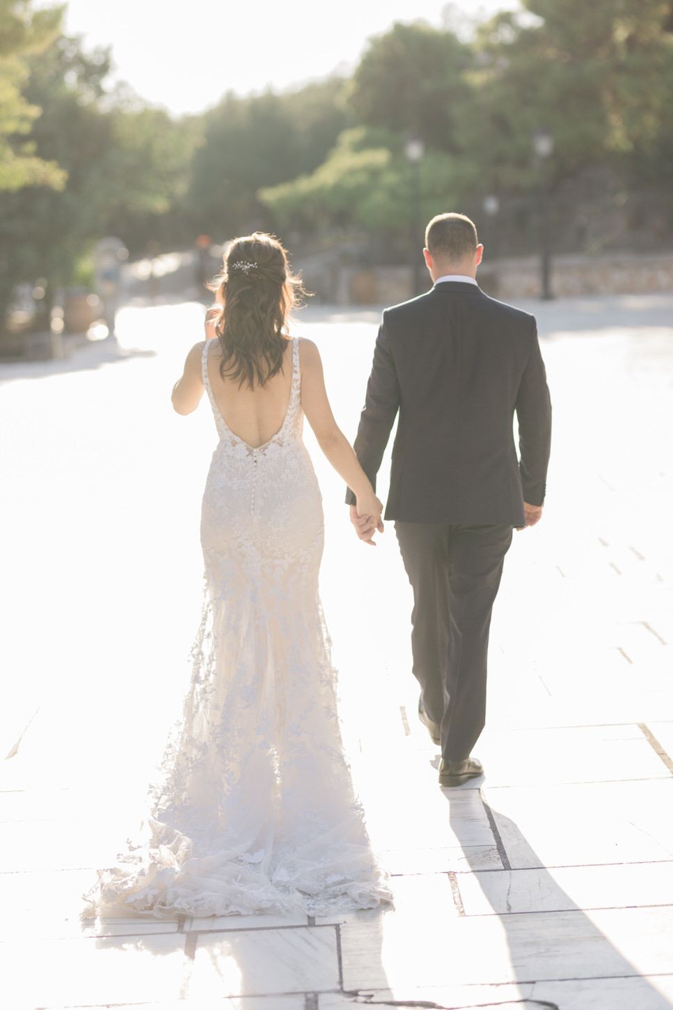 A bride in a lace backless gown and groom in black suit walk hand in hand on a sunlit path.