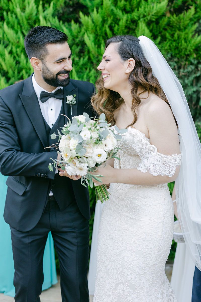A joyful wedding moment captured outdoors with a beautiful white bouquet and elegant formal attire against greenery.