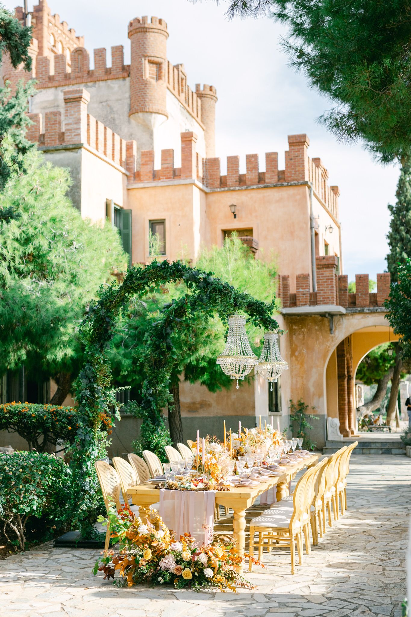 Elegant outdoor wedding table setting with yellow floral arrangements beneath a castle-style villa with crenellated towers.
