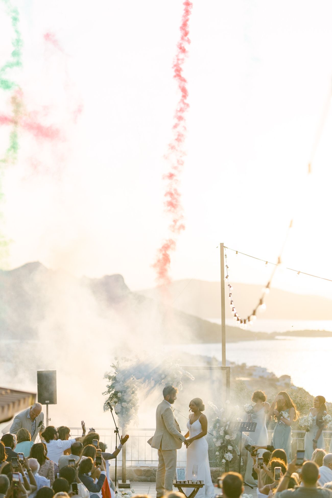 Wedding celebration with colorful smoke effects and crowd of guests during sunset ceremony.
