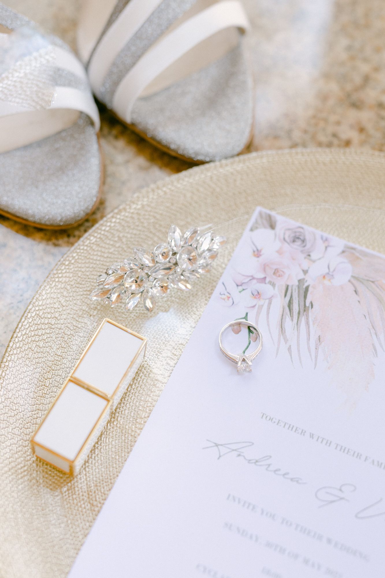 Wedding accessories including silver shoes, jewelry, and invitation card arranged on beige surface.