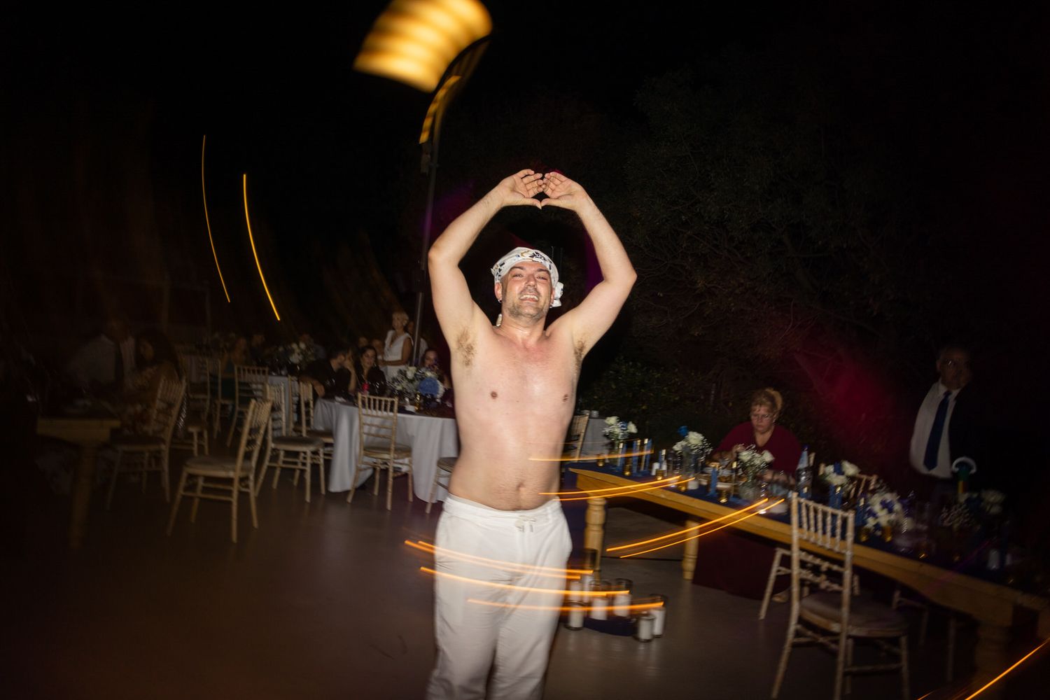 A shirtless performer throws an object above their head during a nighttime indoor performance with tables visible in background.