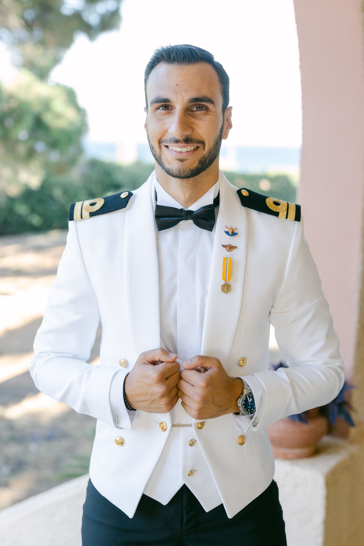 Naval officer in formal white dress uniform with bow tie and gold insignia poses in elegant military attire outdoors.