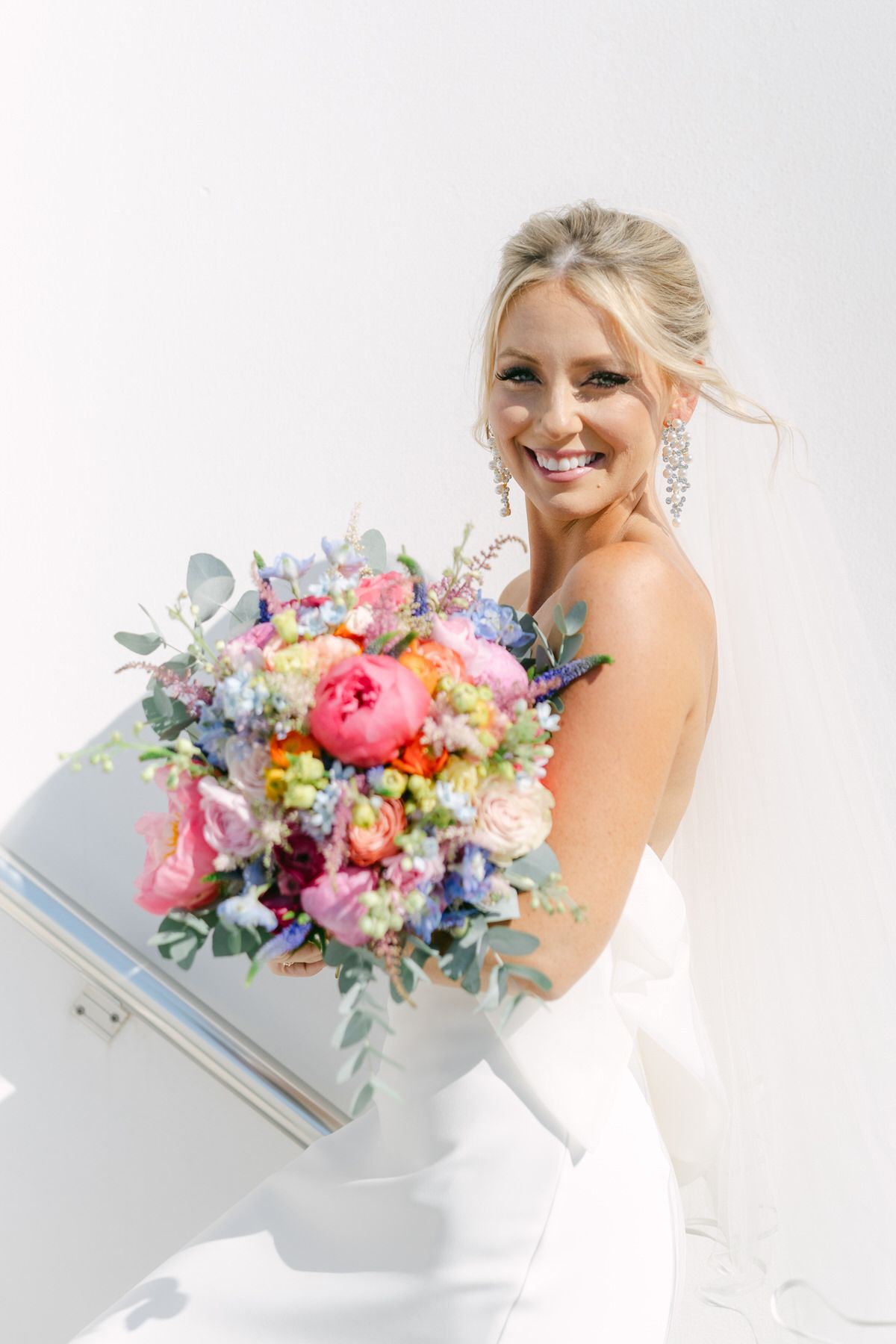 Beautiful bridal portrait showing a white wedding gown and vibrant pink, purple and blue wildflower bouquet against white backdrop.