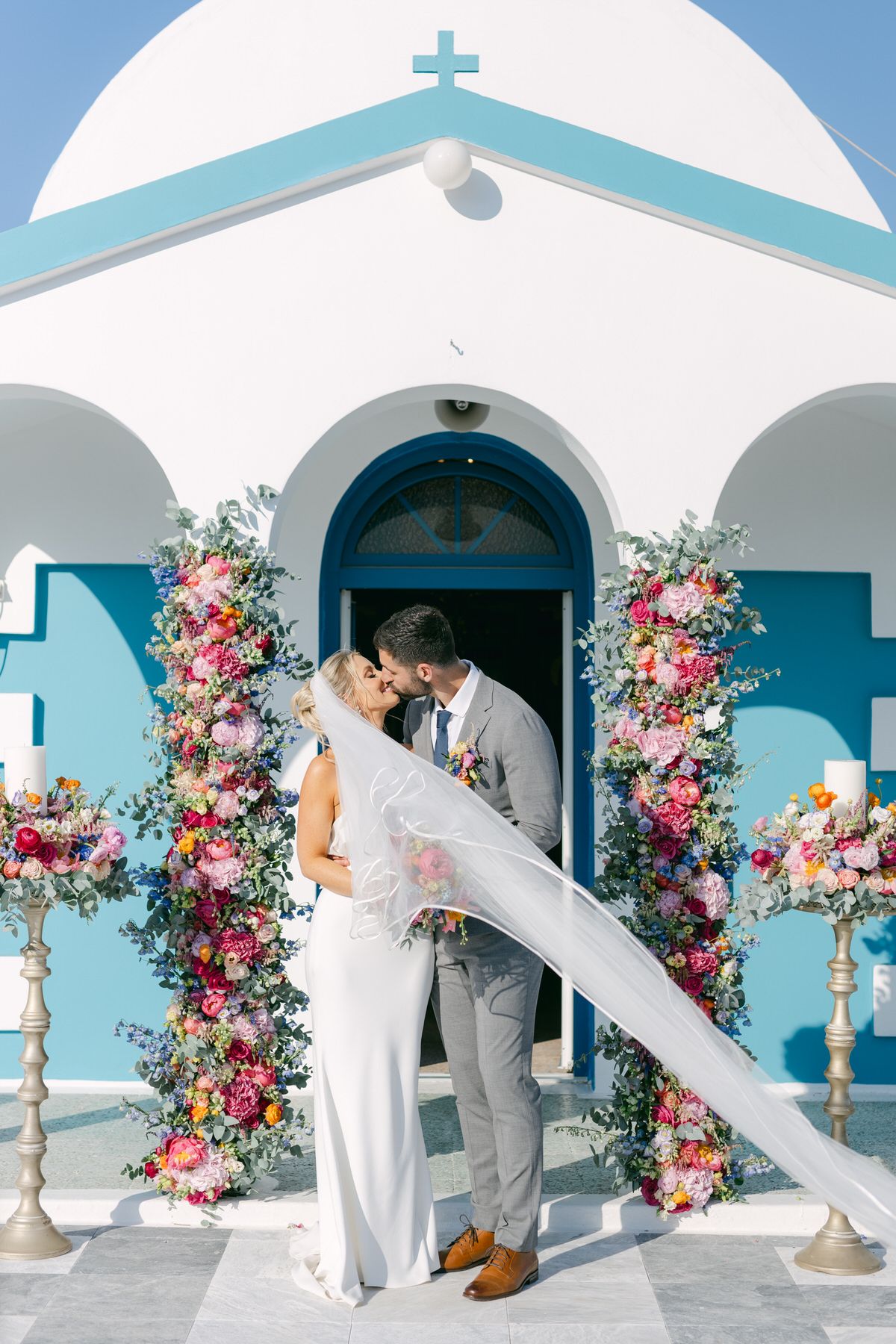 Romantic wedding scene at a blue and white Mediterranean church with vibrant floral arch and flowing veil caught in breeze.