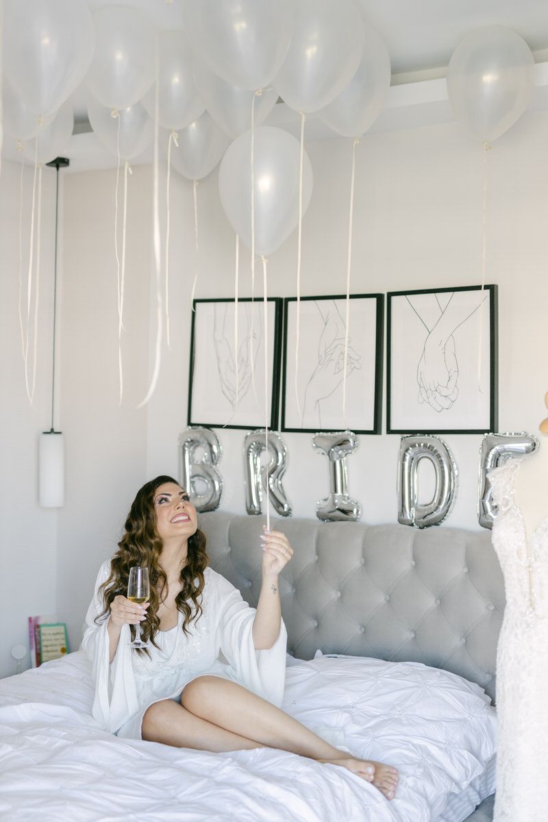 A person in a white robe smiling on a bed with silver BRIDE balloons and white decorations.