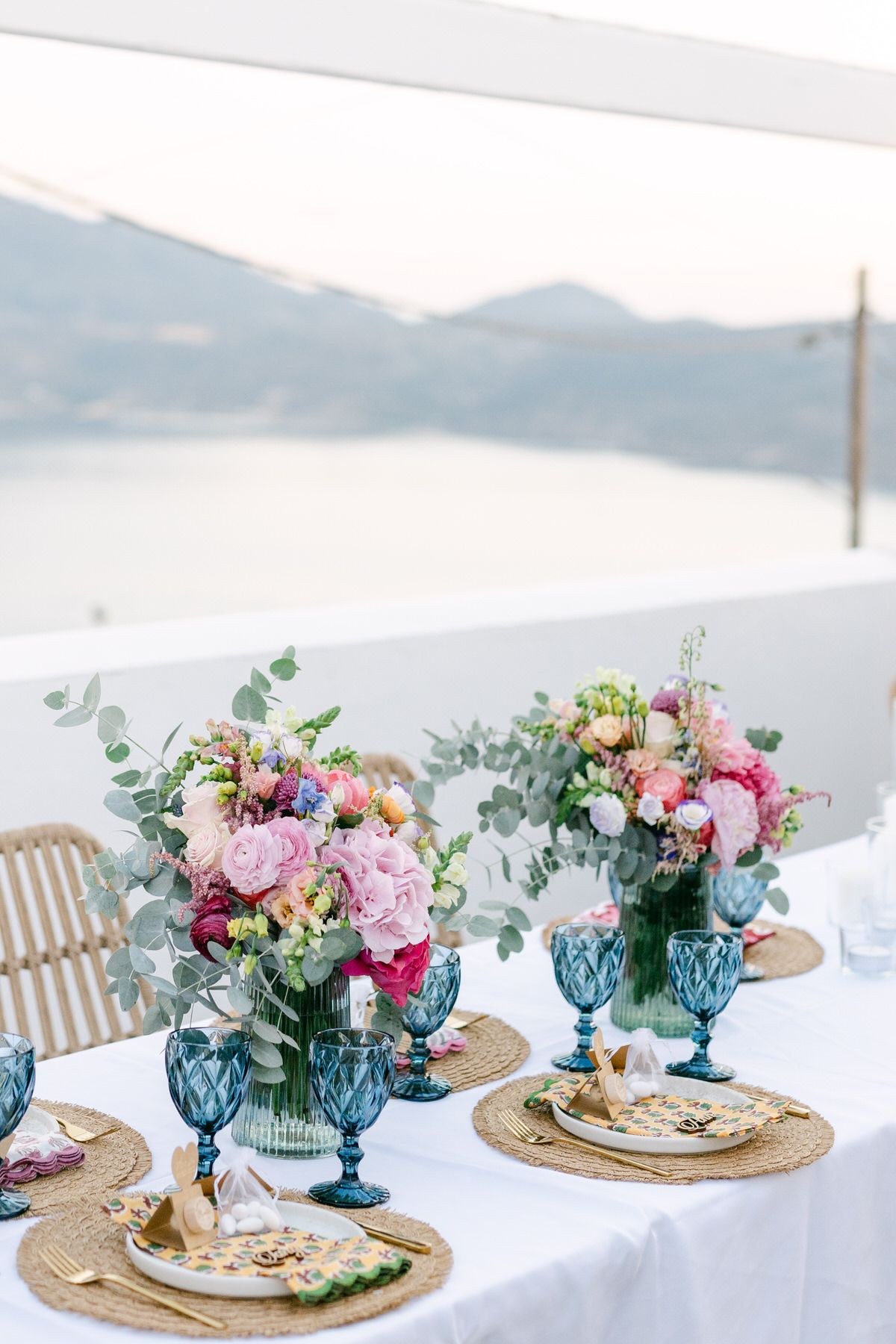 Elegant table setting with colorful floral centerpieces, blue goblets, and gold chargers overlooking a misty mountain view.