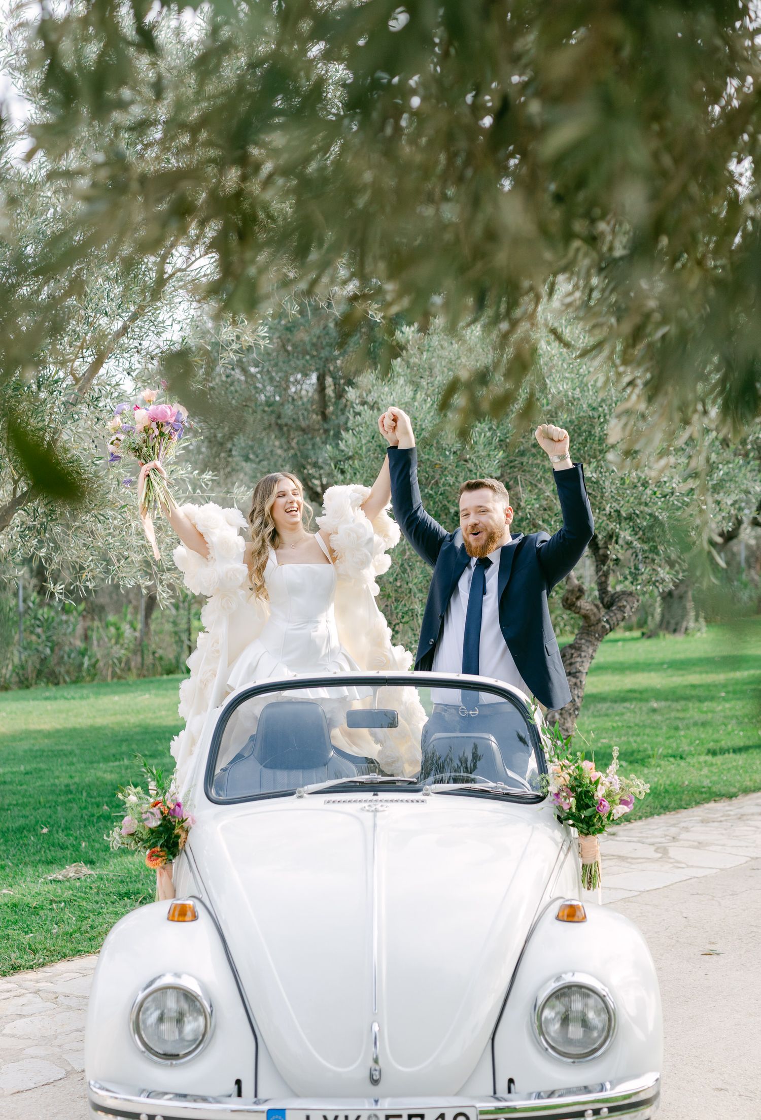 A joyful wedding couple celebrates in a classic white Volkswagen Beetle decorated with flowers in a garden setting.
