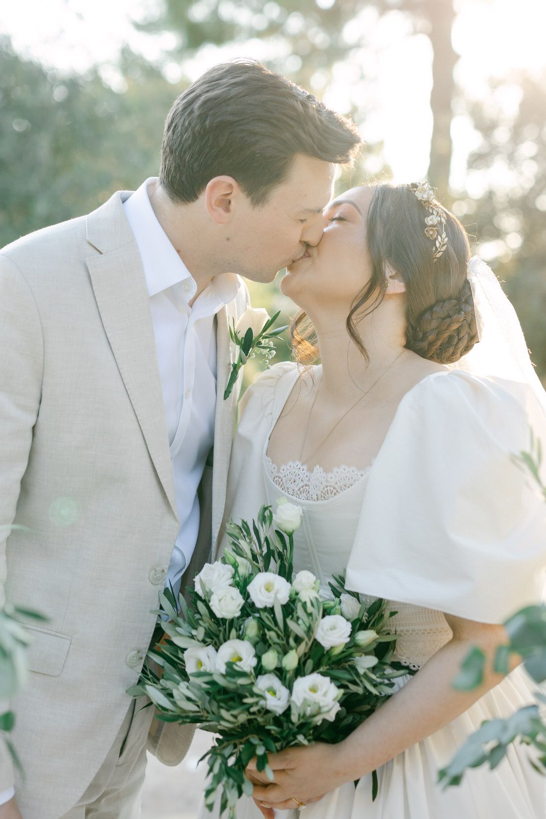 A bride and groom share a romantic kiss while holding a white floral bouquet in an outdoor garden setting at sunset.
