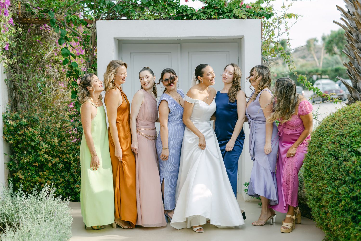 A bridal party wearing colorful pastel bridesmaid dresses in different styles poses together in a garden archway.