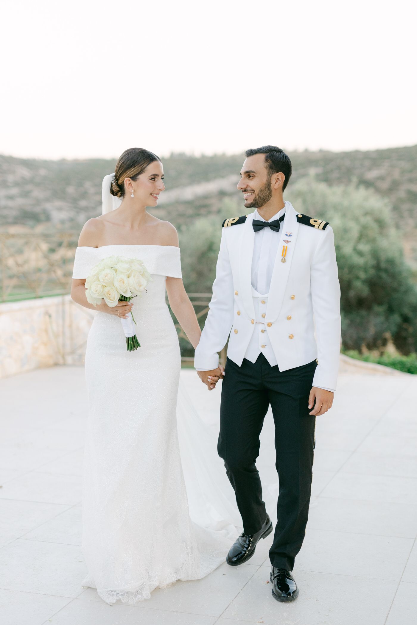 A couple in wedding attire walks hand in hand, with the bride in an off-shoulder gown and groom in a white military uniform.