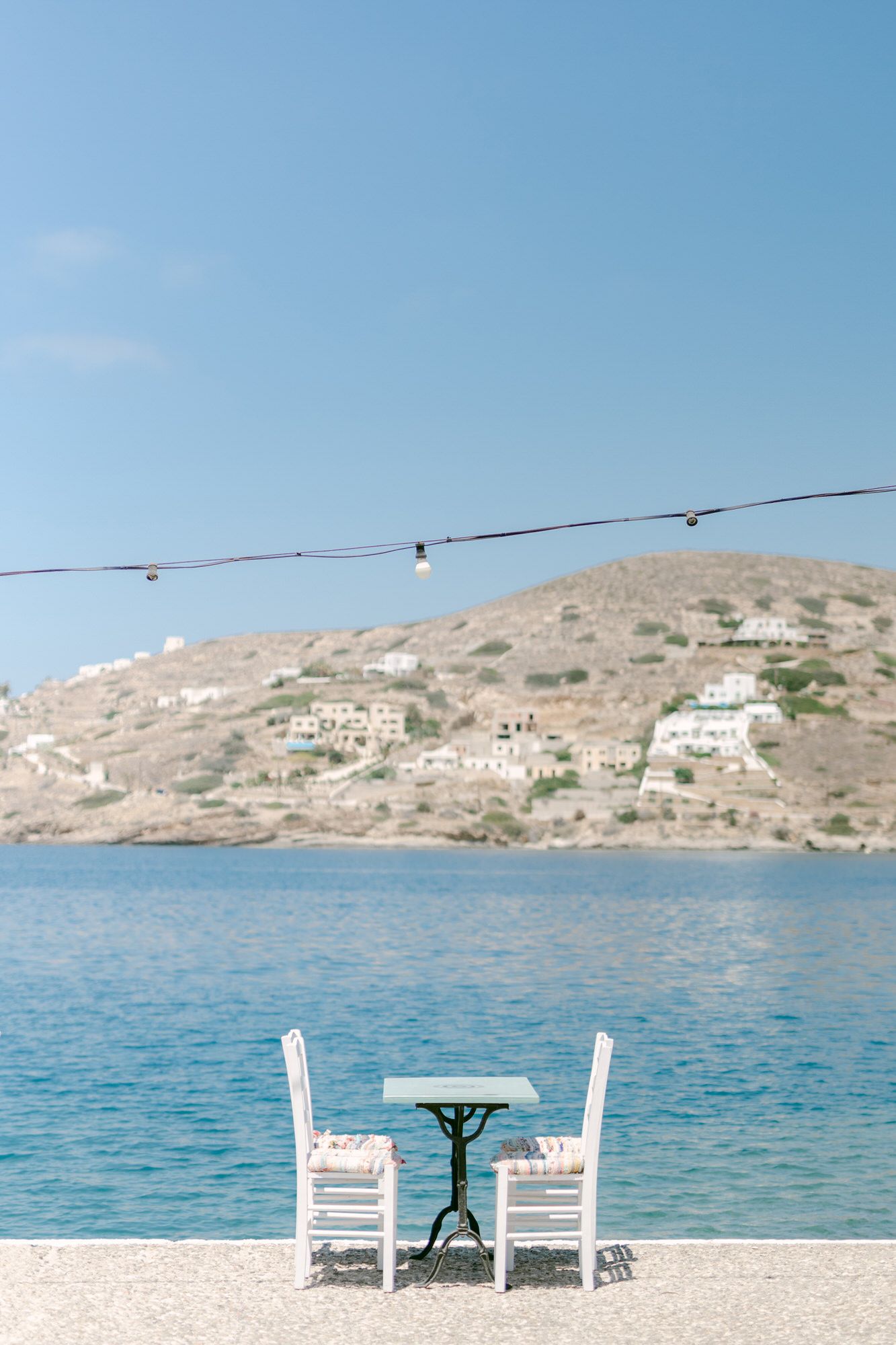 Two white chairs and small table overlooking the Mediterranean Sea with coastal homes on hillside.