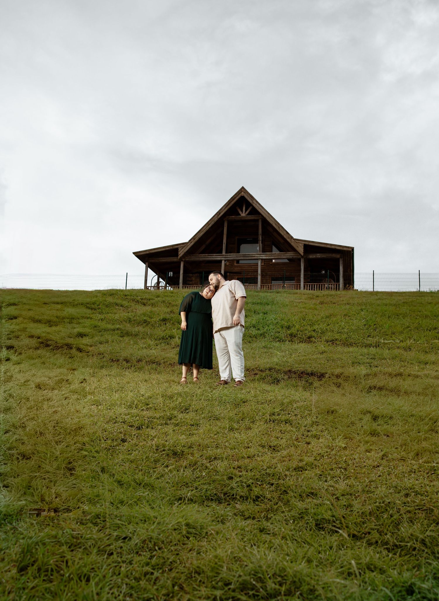 Rustic wooden cabin sits atop a green hillside under cloudy skies.