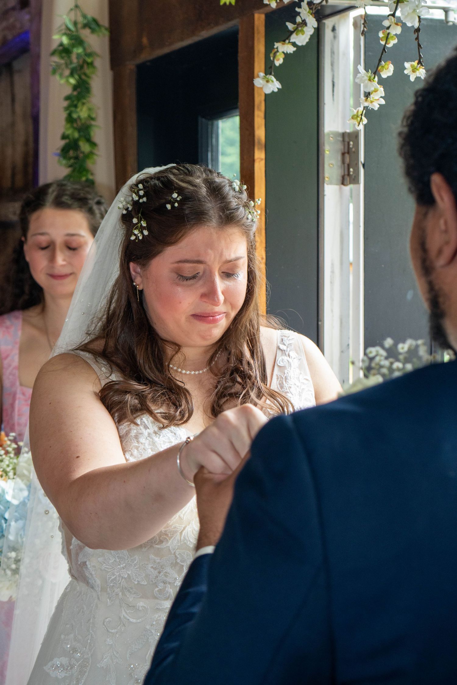 A tender moment between newlyweds in white dress and navy suit near a window with floral decorations.