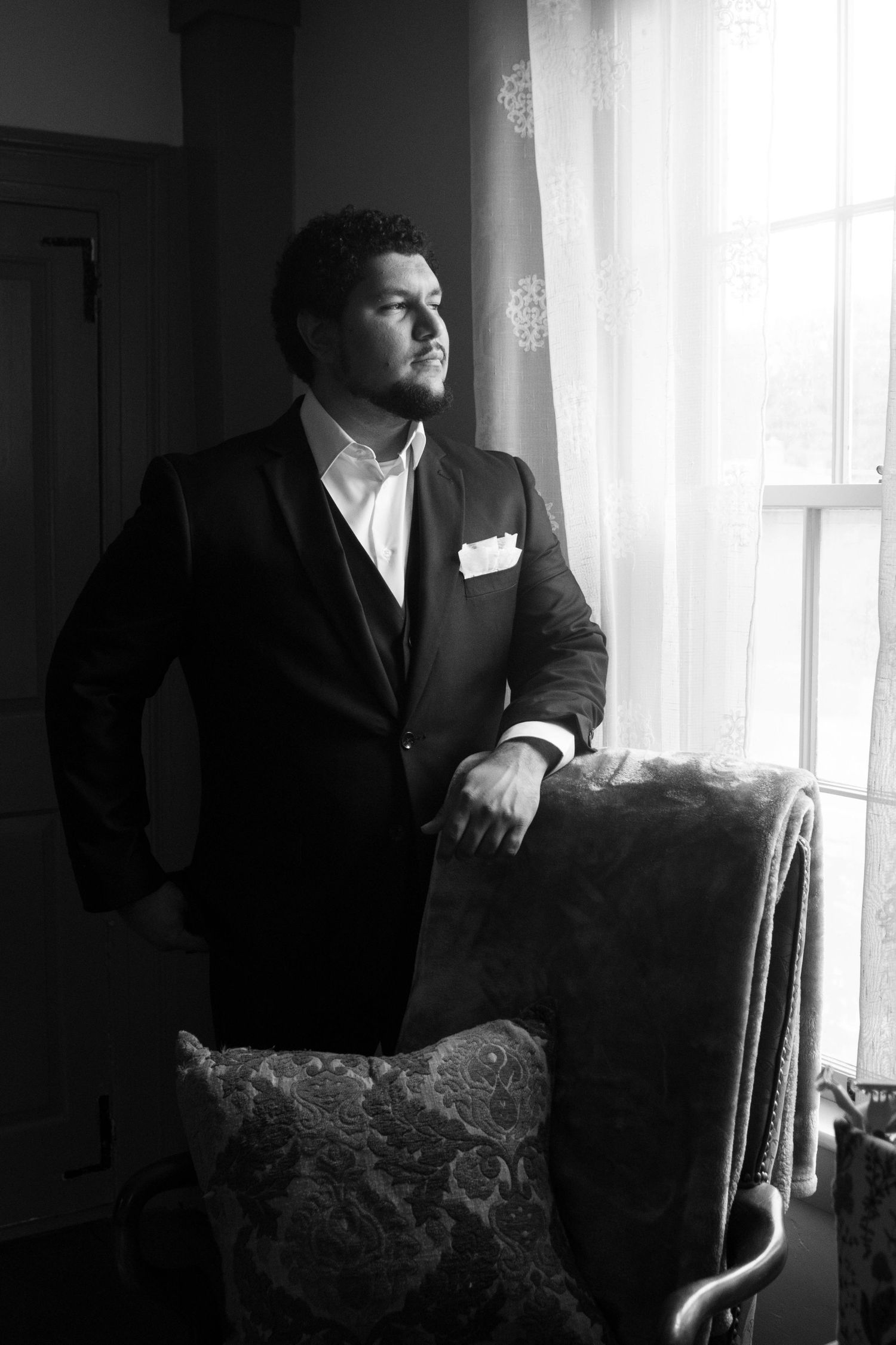 Dramatic black and white portrait by window with moody lighting and vintage chair.