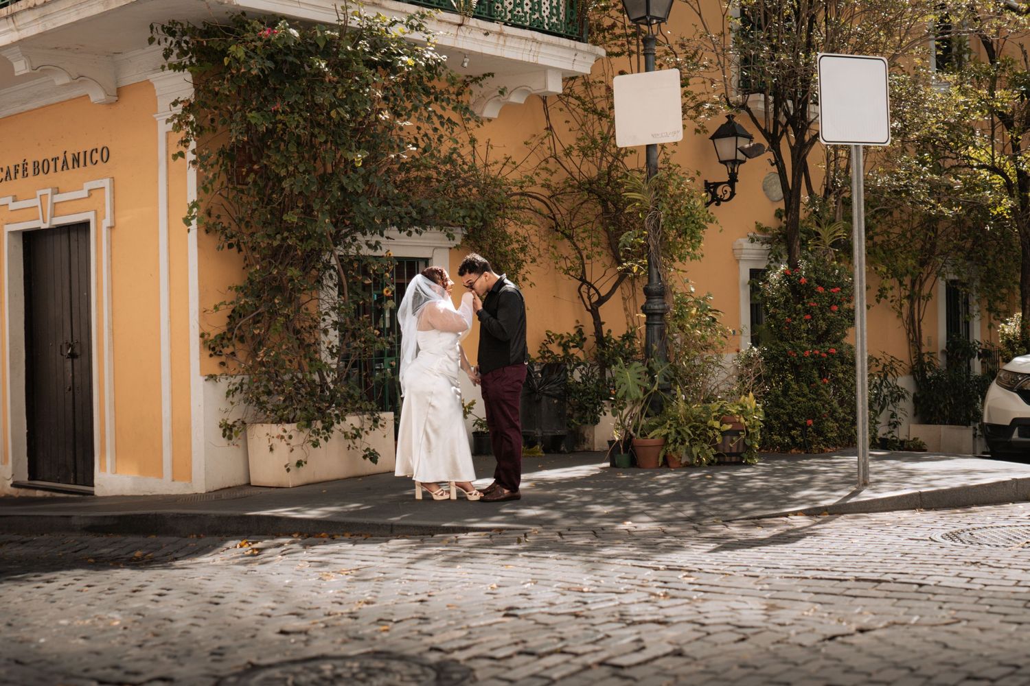 A wedding couple walks along a sunlit path beside a yellow building with trees.