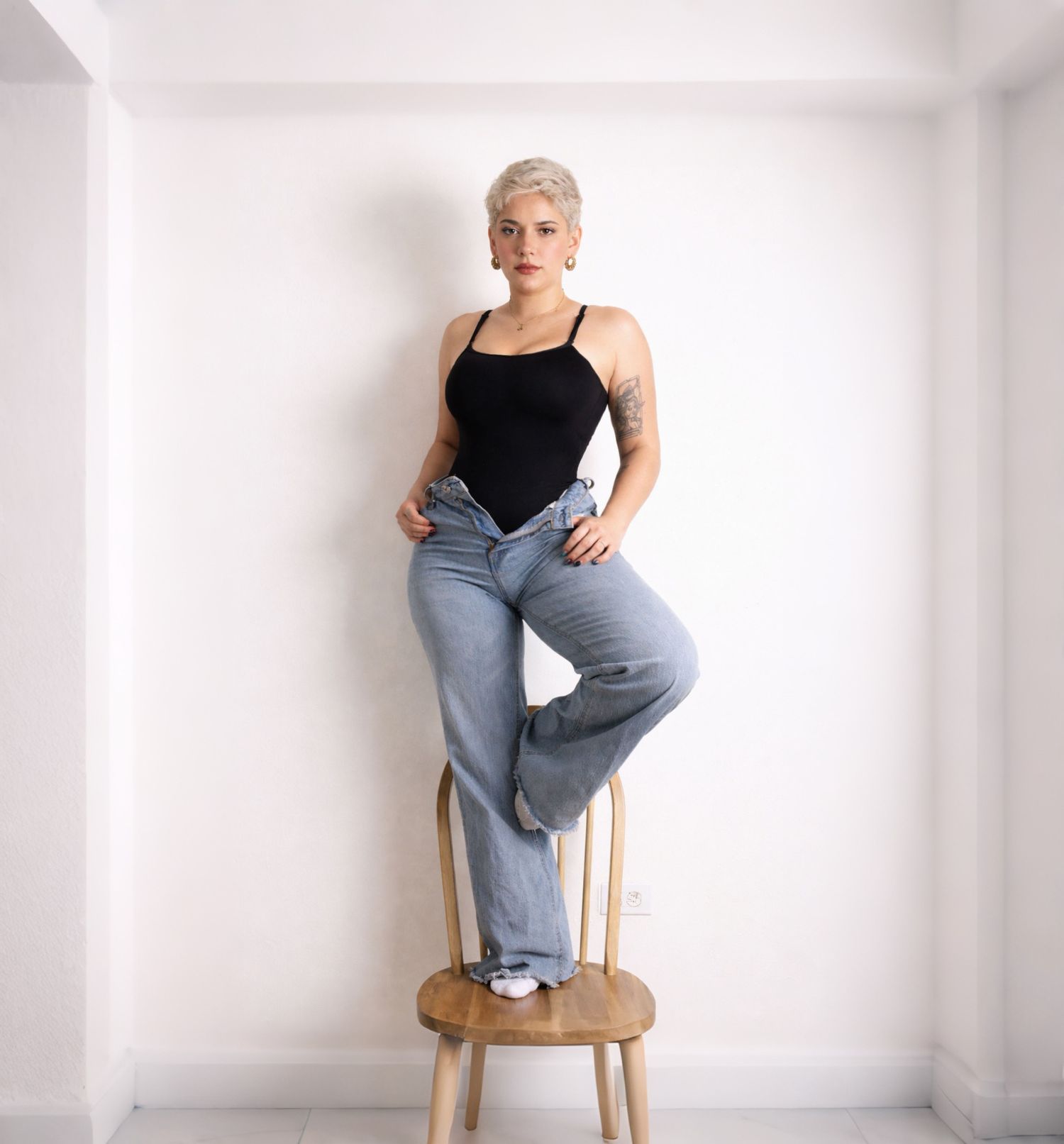 A person in a black tank top and jeans sits casually on a wooden chair against a white wall.