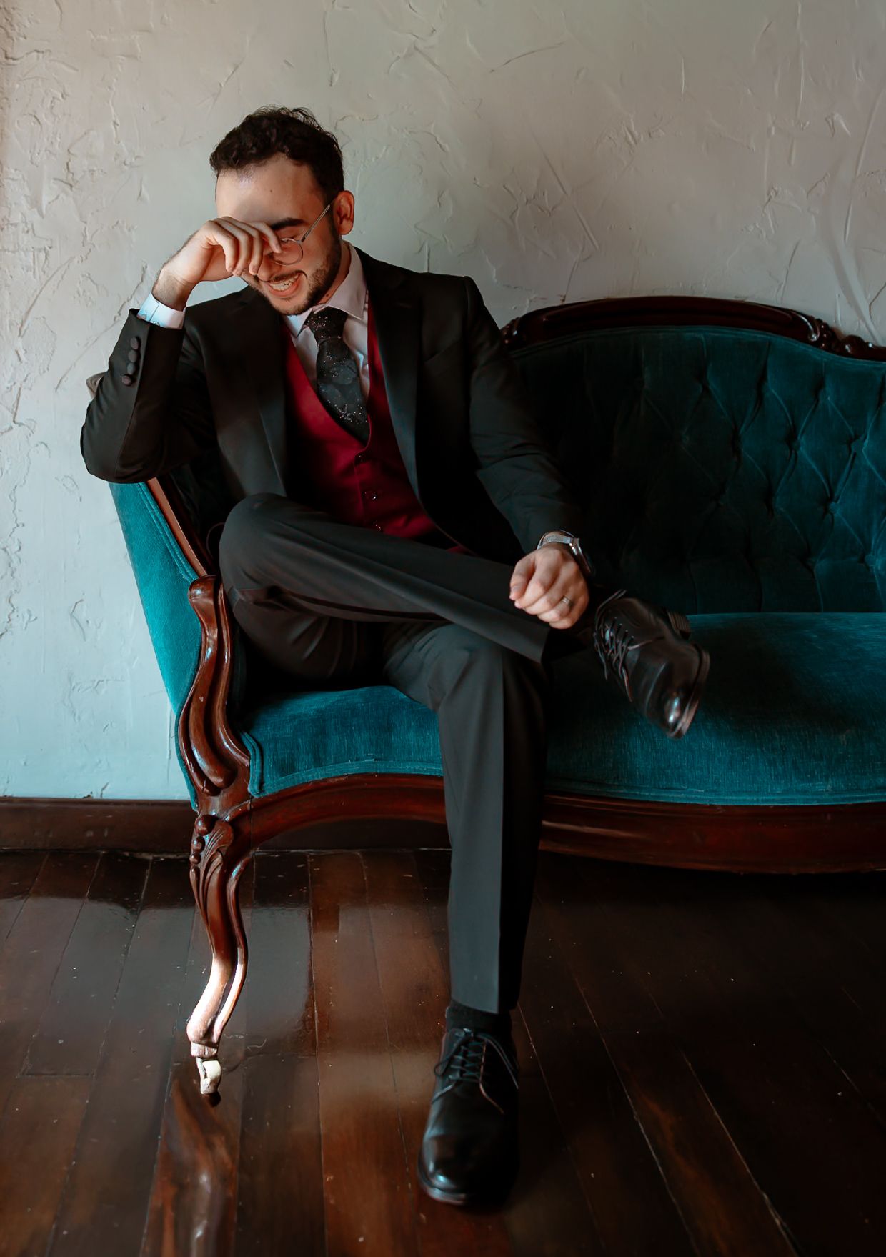 Groom in black suit and red tie sits pensively on vintage teal sofa with hardwood floors.