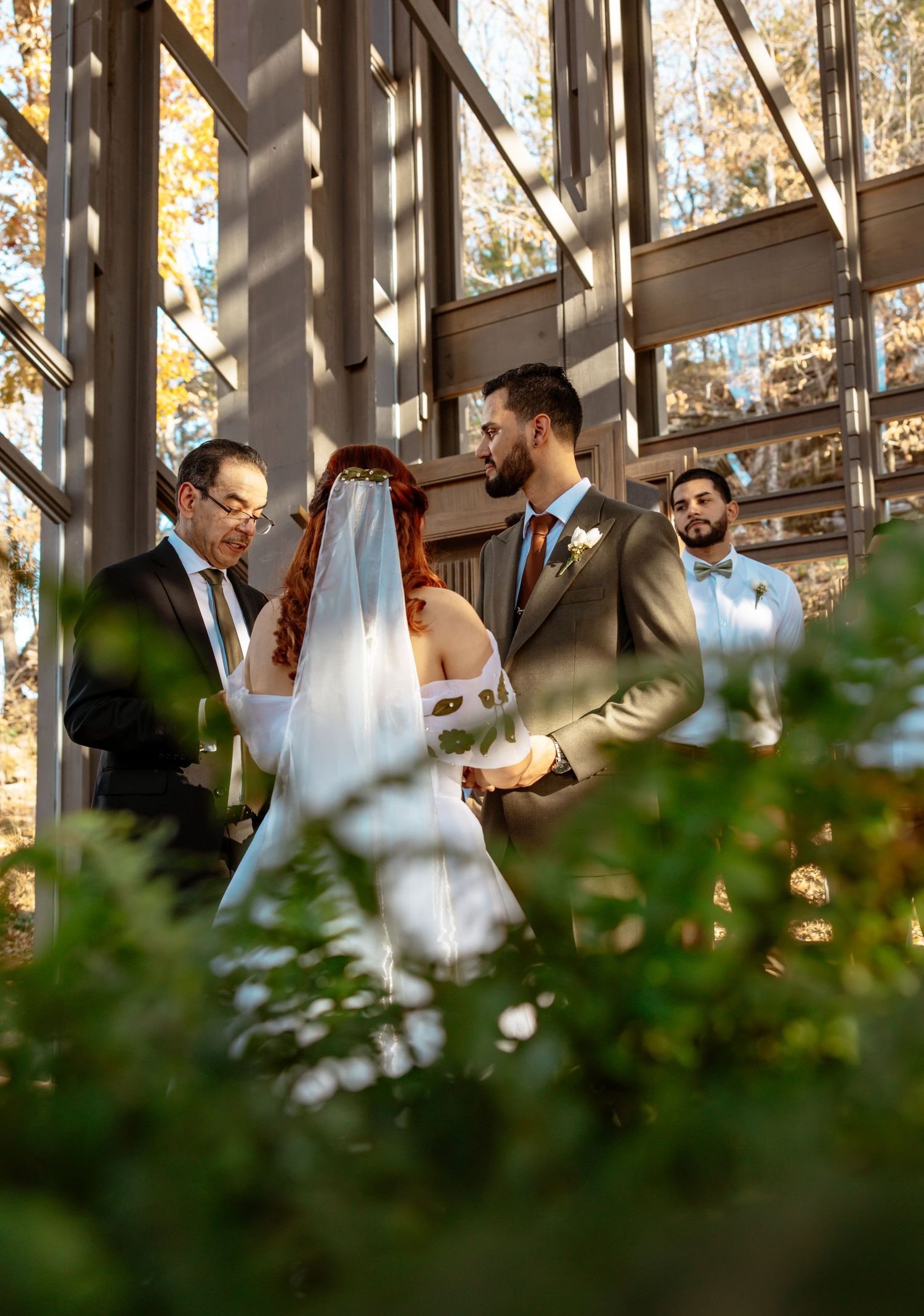 A wedding ceremony takes place in a sunlit glass-walled venue surrounded by autumn trees and greenery.