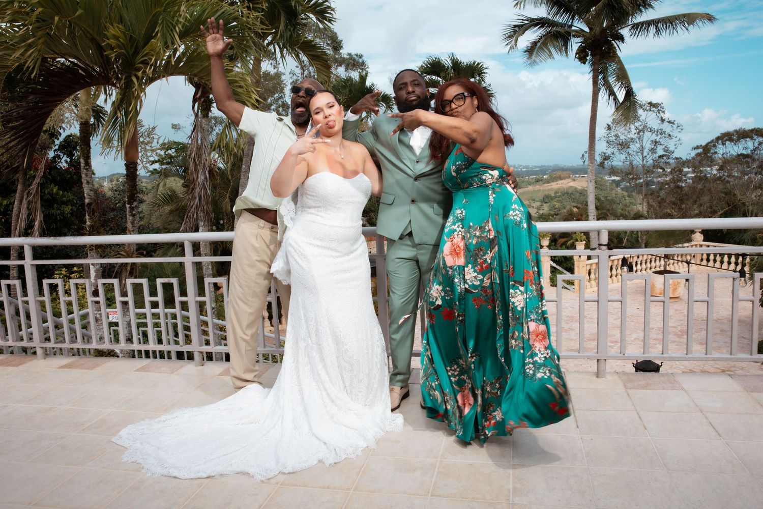 A couple in wedding attire poses together on a beachfront deck with palm trees swaying in the background.