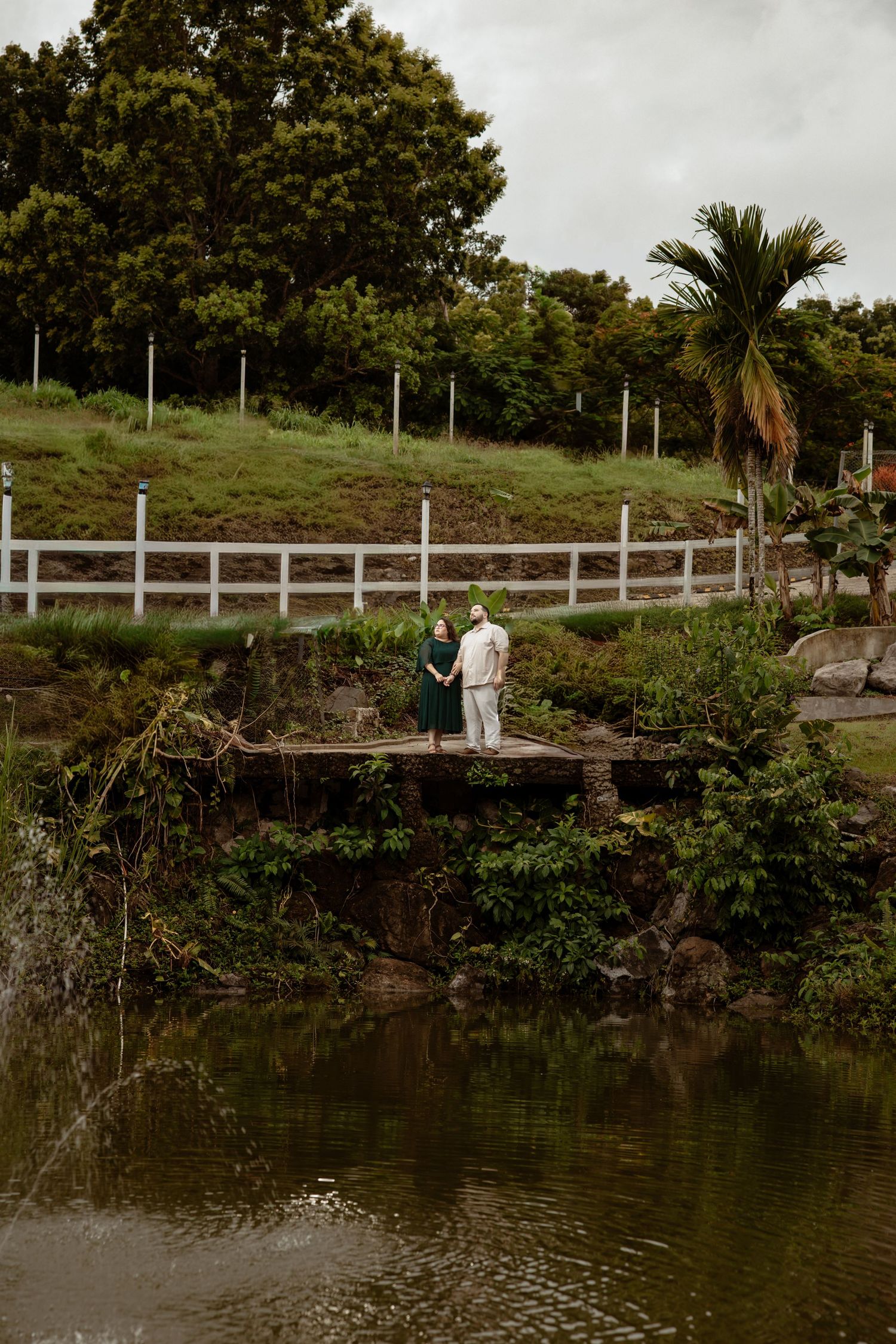 A couple stands on a wooden bridge overlooking a serene pond with palm trees.