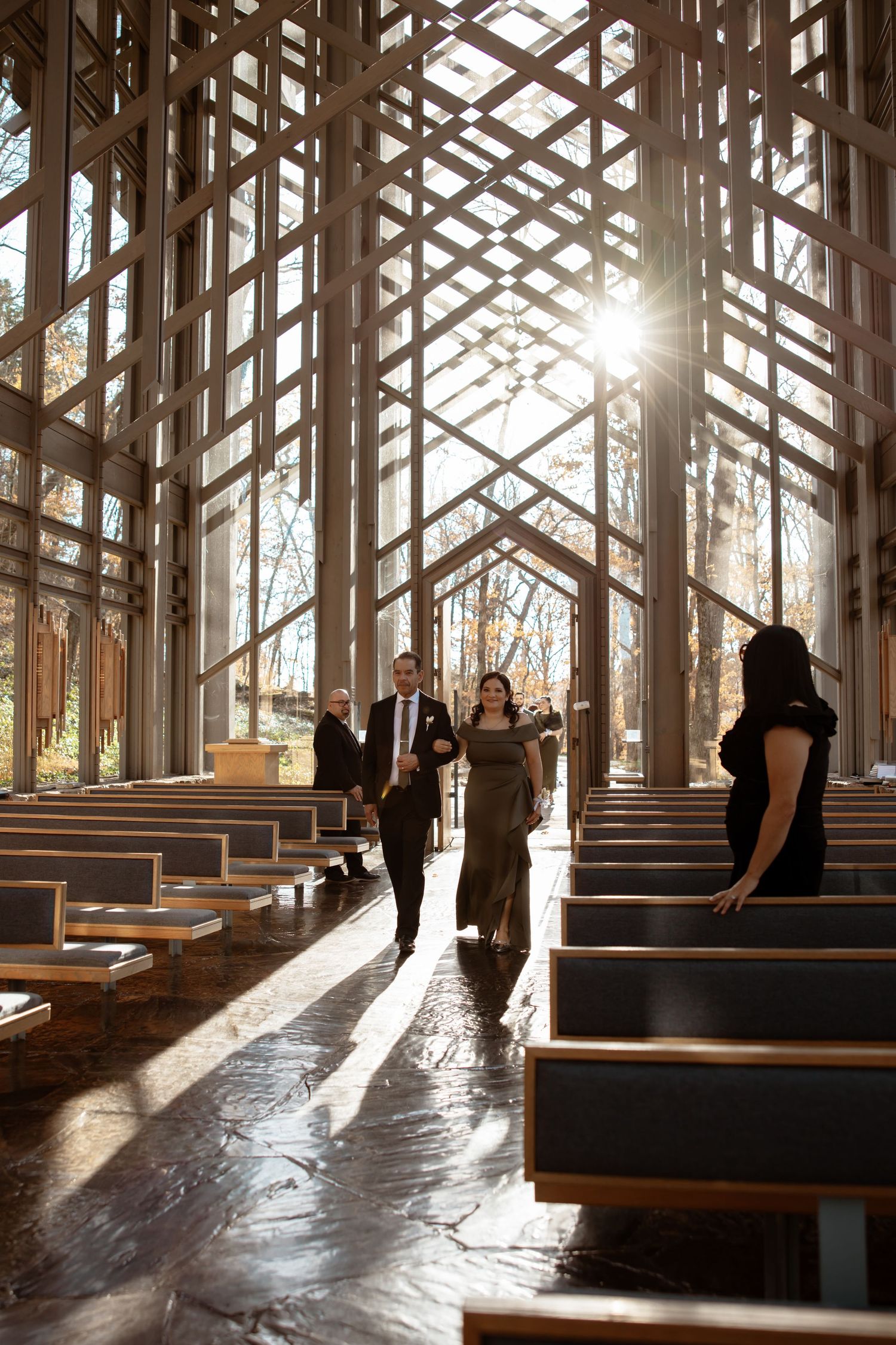 Silhouettes of people walk through a sunlit wooden chapel interior with dramatic shadows and geometric patterns.