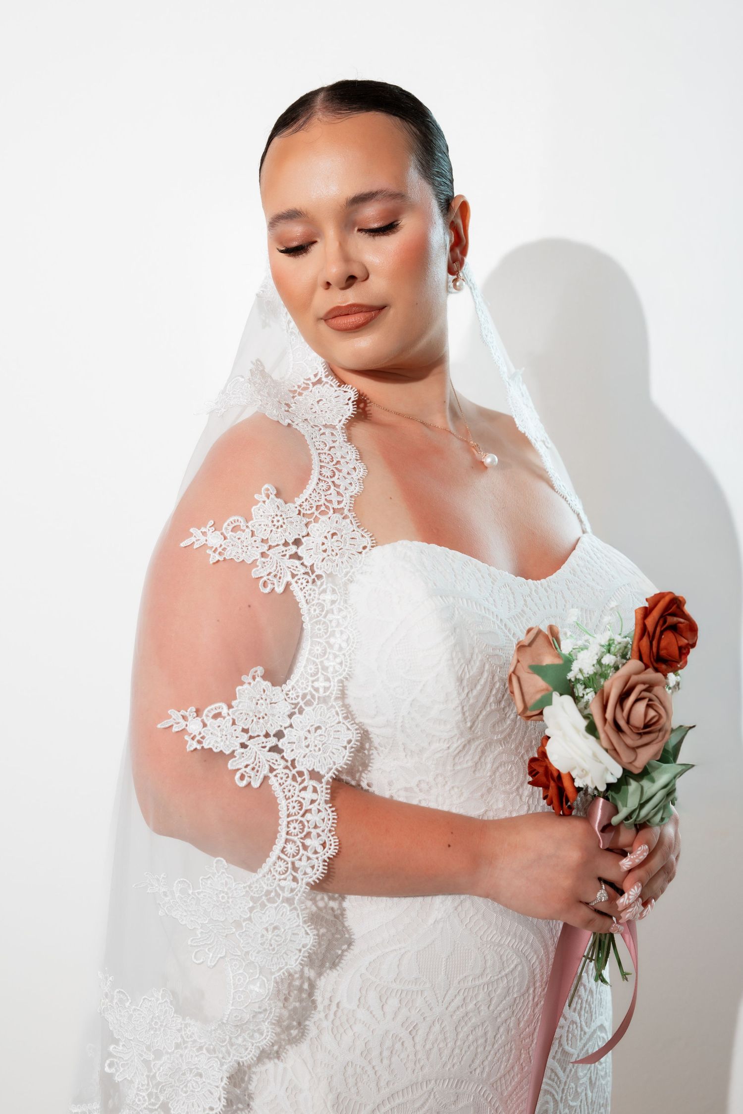 A bride in a white lace wedding dress with sheer sleeves holds burgundy and brown roses against a white background.