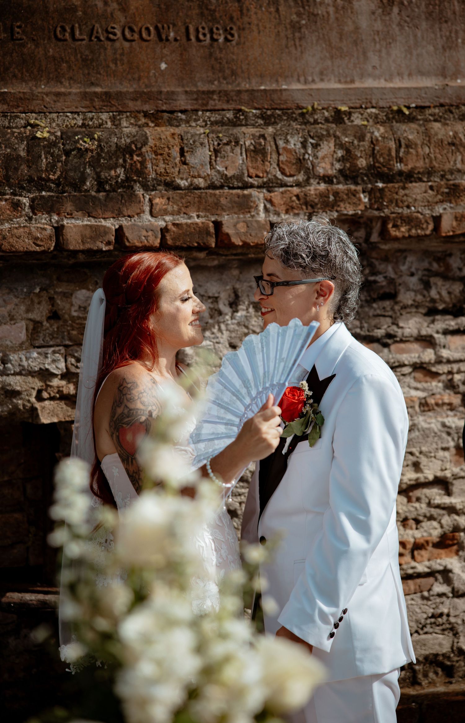 Wedding couple shares intimate moments against a rustic brick wall with white flowers in foreground.