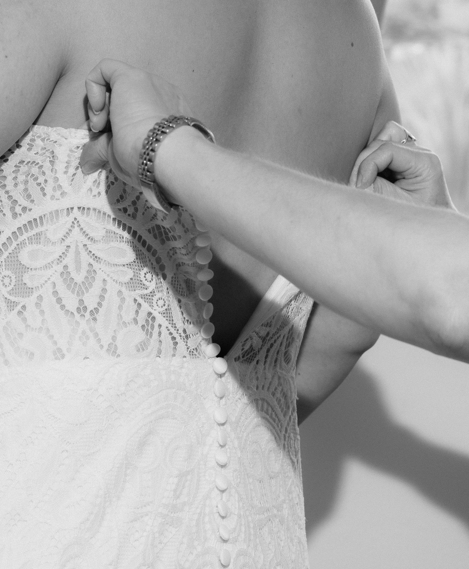 Close-up view of a bride's white lace wedding dress being fastened with delicate buttons down the back.